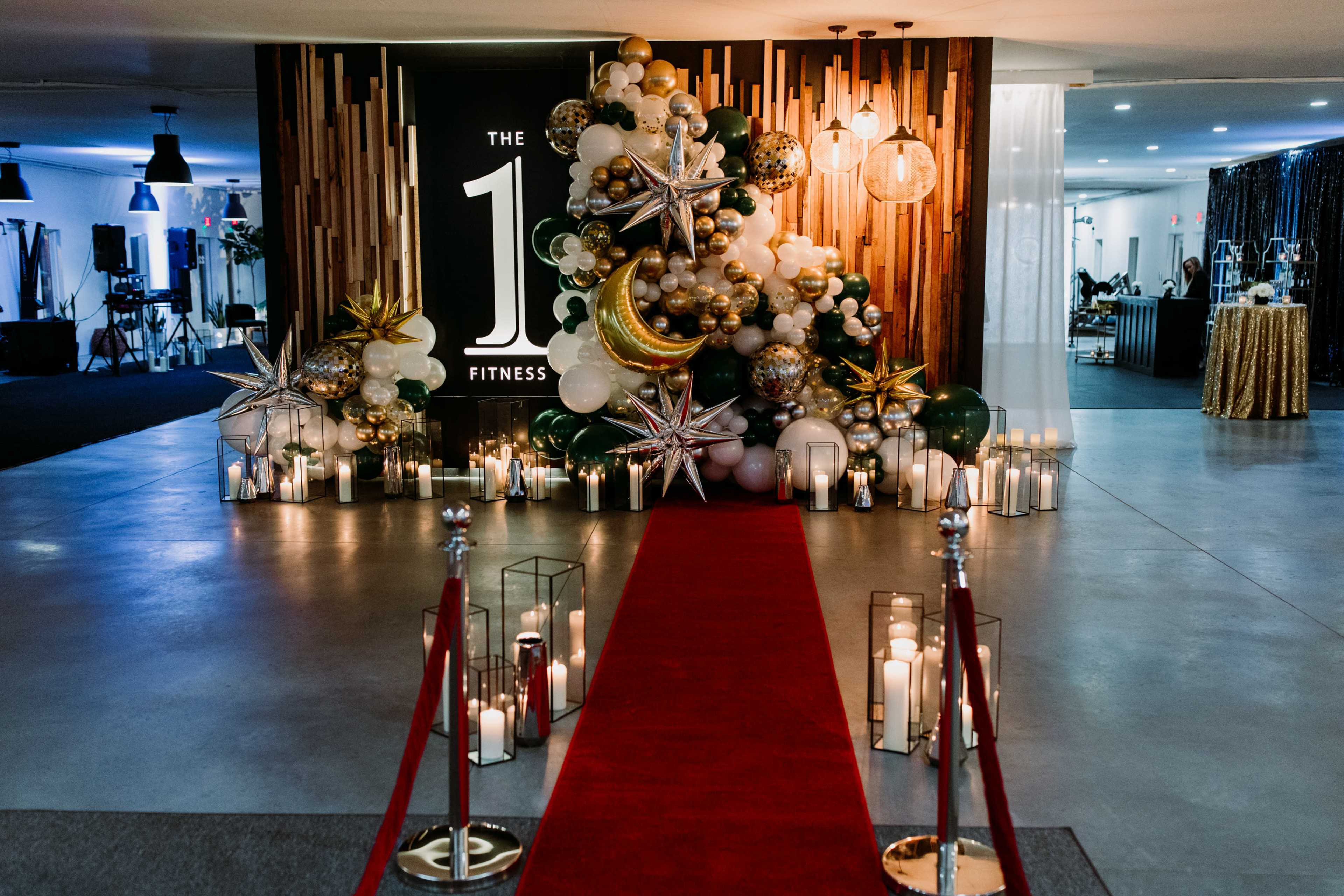 A red carpet leads to a decorative display of balloons and lights beside a sign that reads "THE 1 FITNESS."