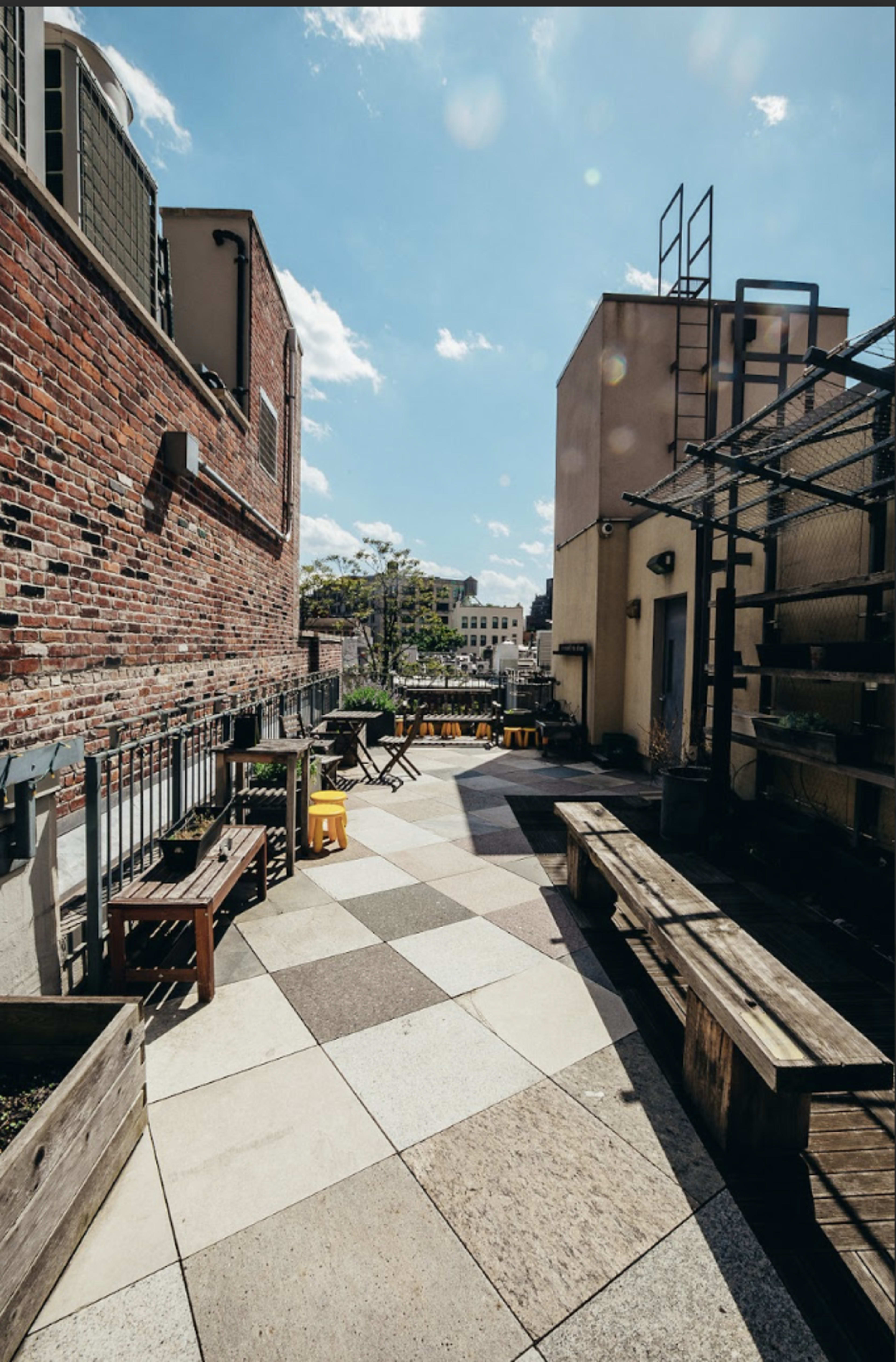 The image shows a rooftop terrace with a patterned stone floor, wooden benches, and planters under a bright blue sky with a few clouds.