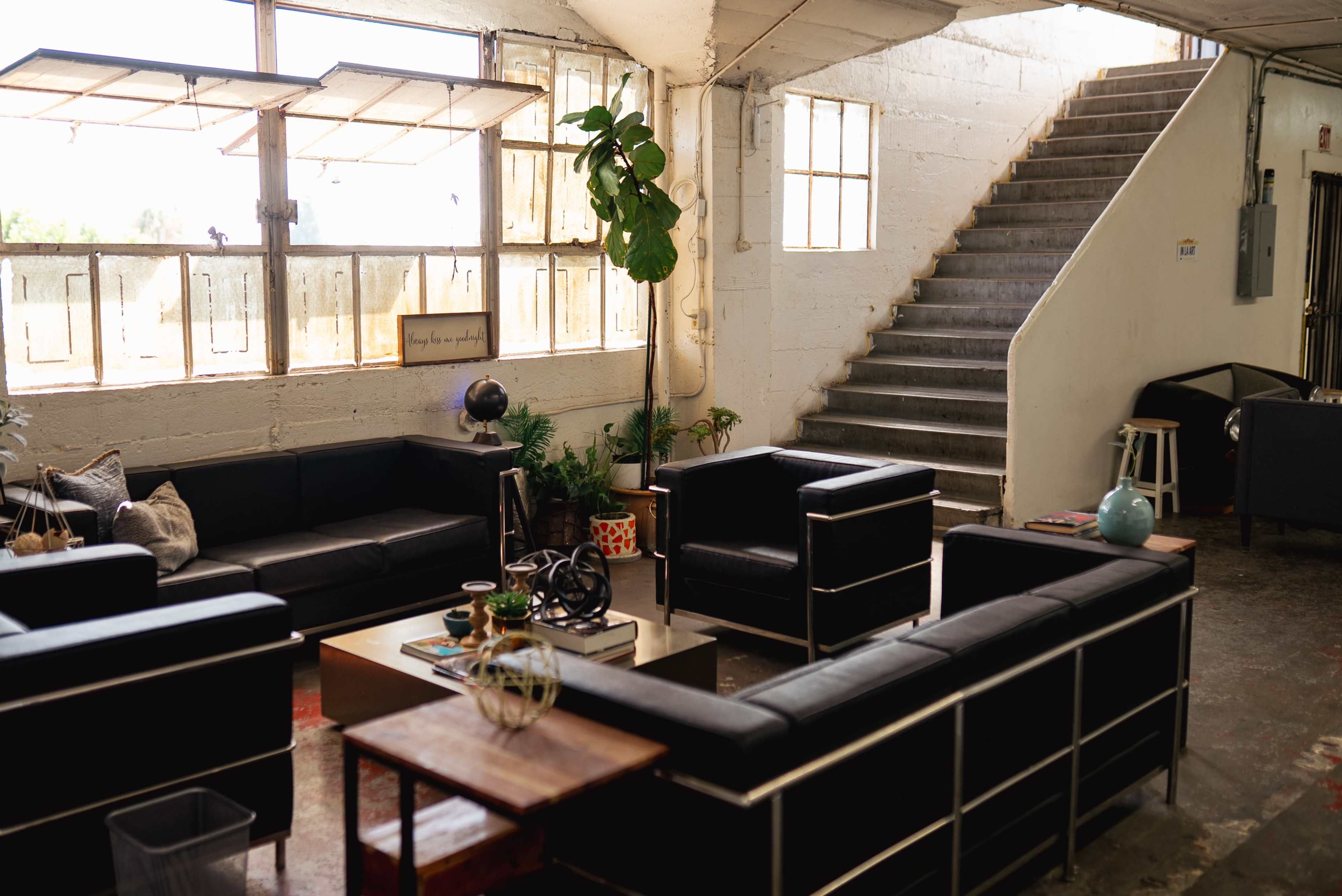 The image shows a spacious, industrial-style indoor area featuring black leather couches arranged around a coffee table, with large windows allowing natural light to flood the space and a staircase leading to an upper level.
