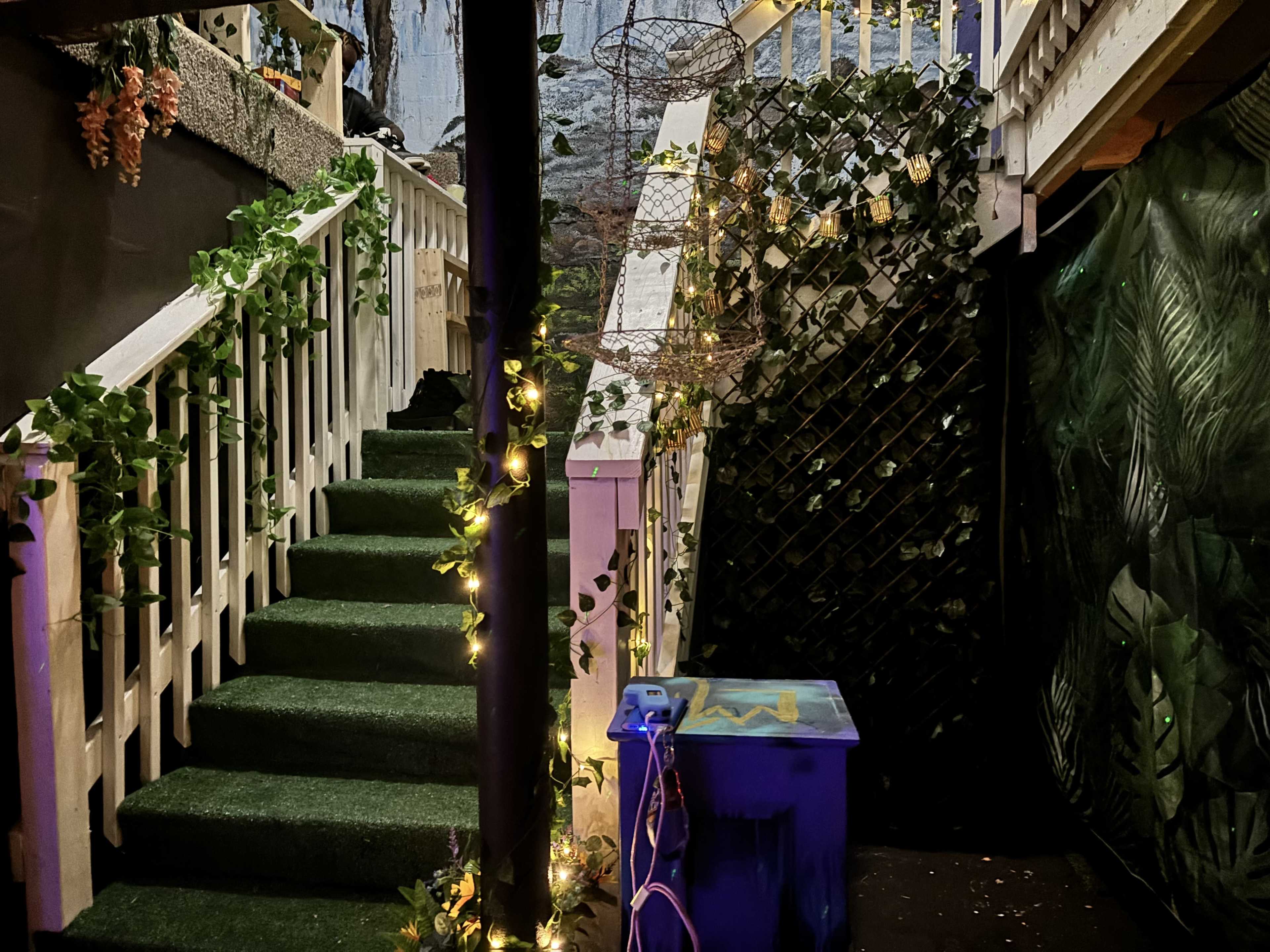 The image shows a staircase with green carpeting leading up, flanked by decorative plants and string lights, set against a backdrop of artificial foliage.