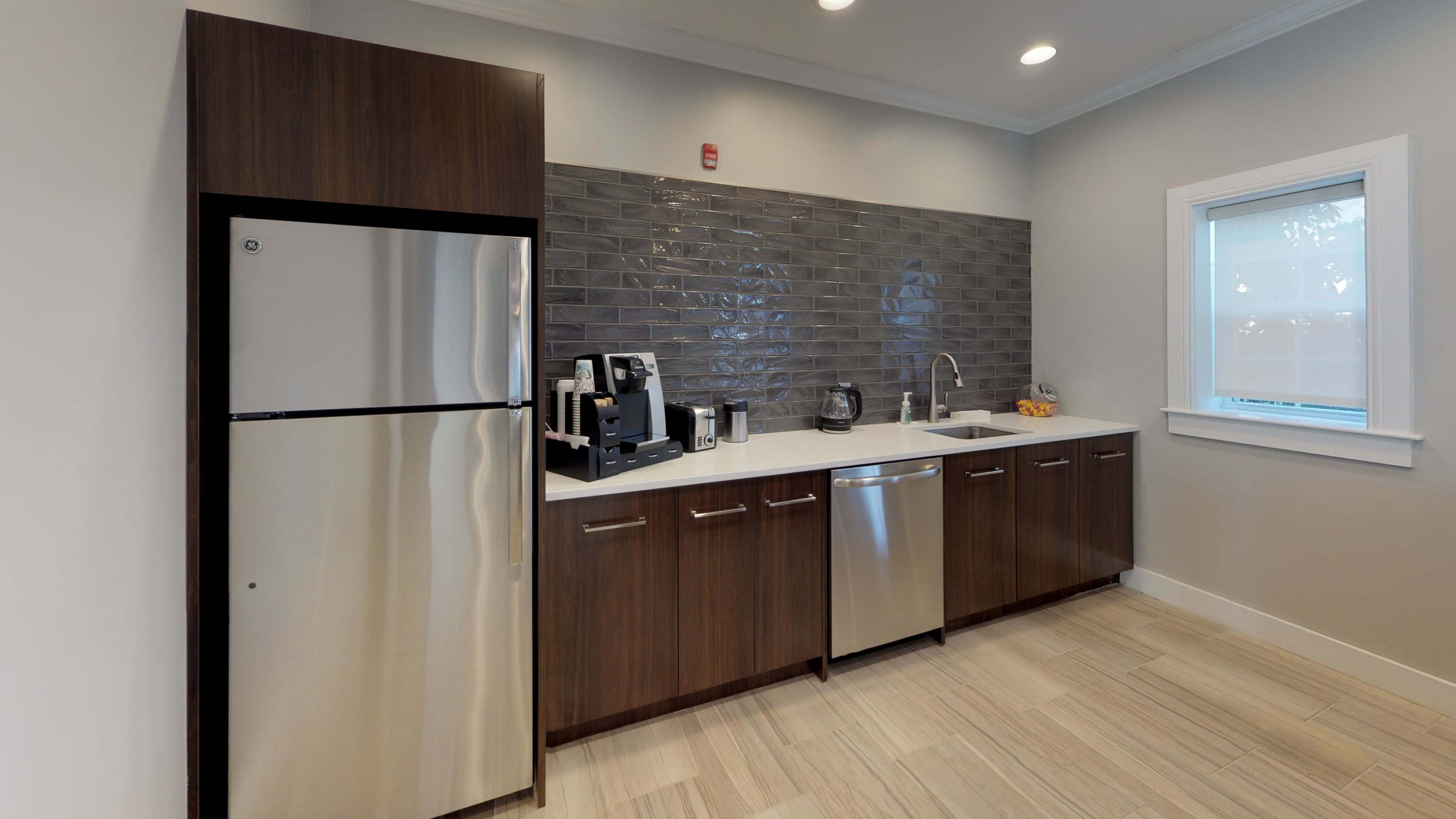 A modern kitchen with a stainless steel refrigerator, dark wood cabinetry, a sink, and a countertop equipped with a coffee maker and other appliances.