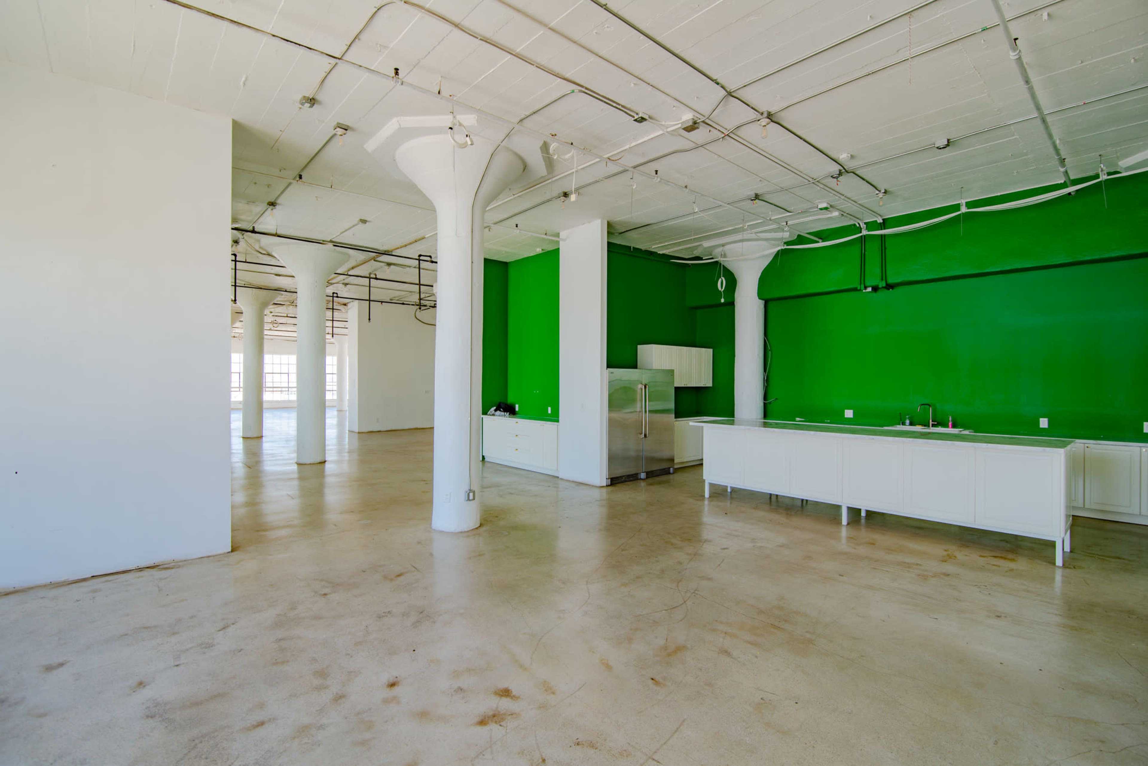 A spacious, empty room with polished concrete floors, white walls, and one vibrant green accent wall, featuring a kitchen area with white cabinetry.