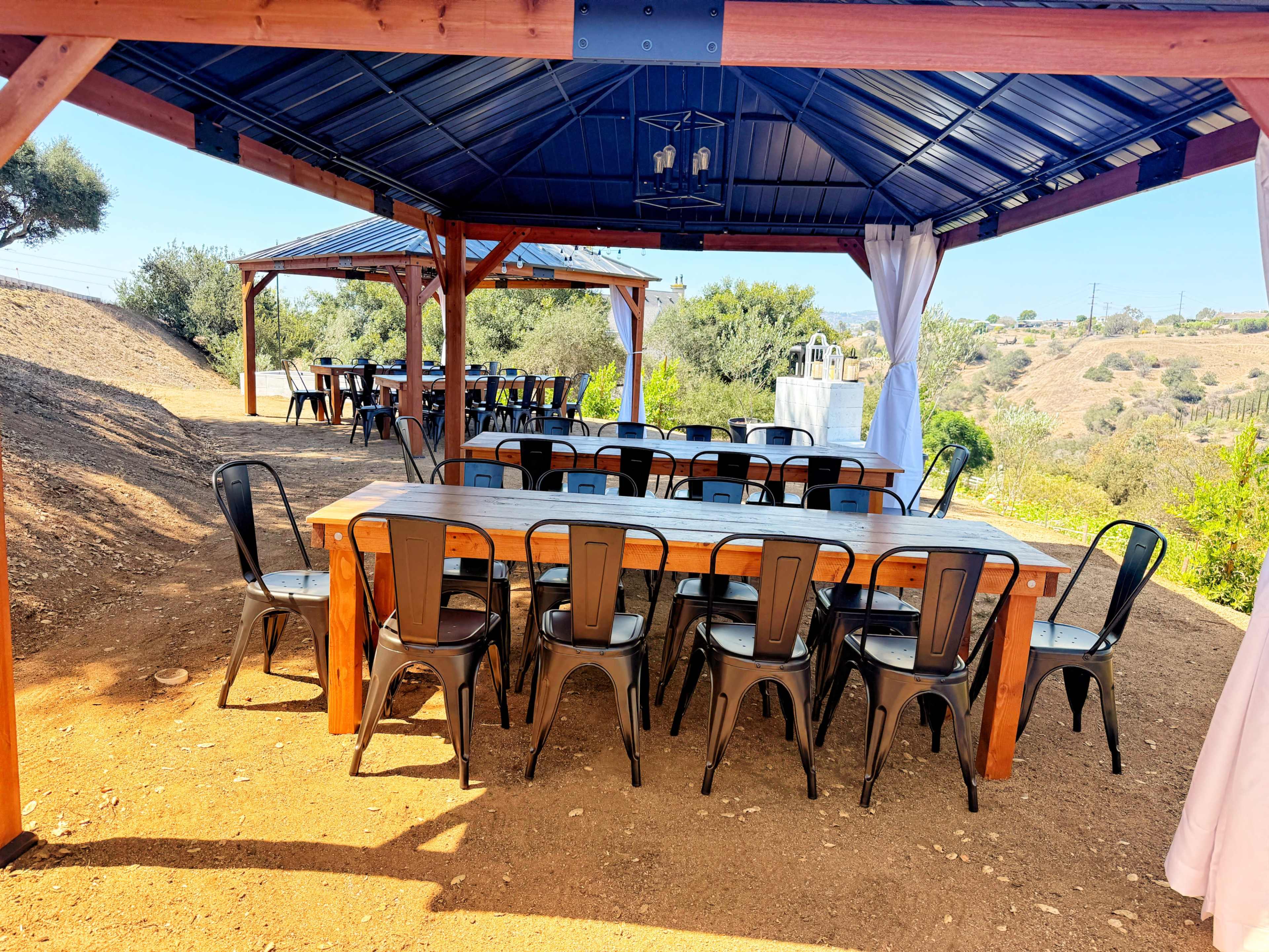 A large wooden table with metal chairs is set up under a wooden gazebo on a hillside surrounded by greenery.