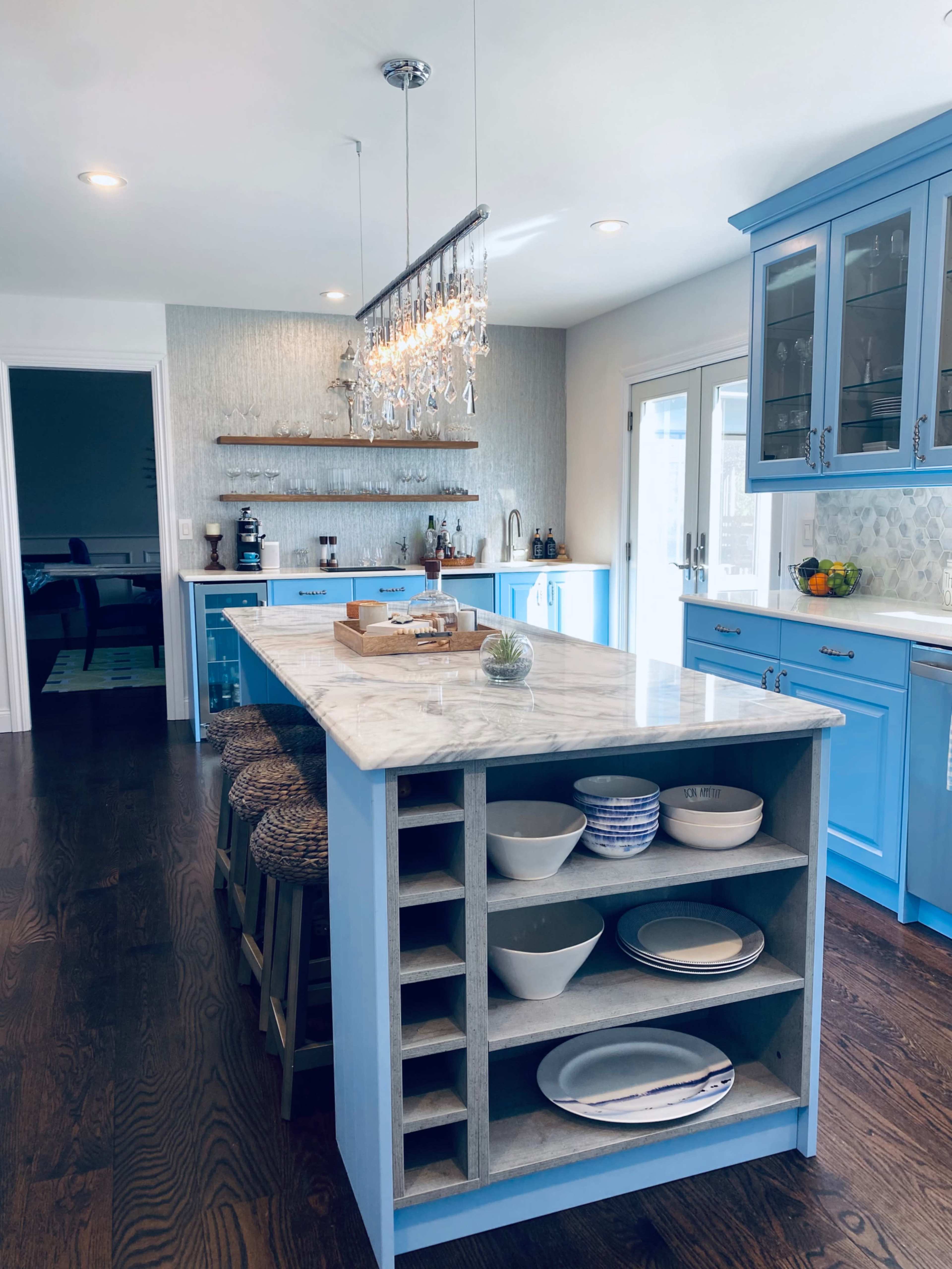 A modern kitchen features a blue island with a marble countertop, surrounded by wooden stools, sleek cabinetry, and a chandelier overhead.