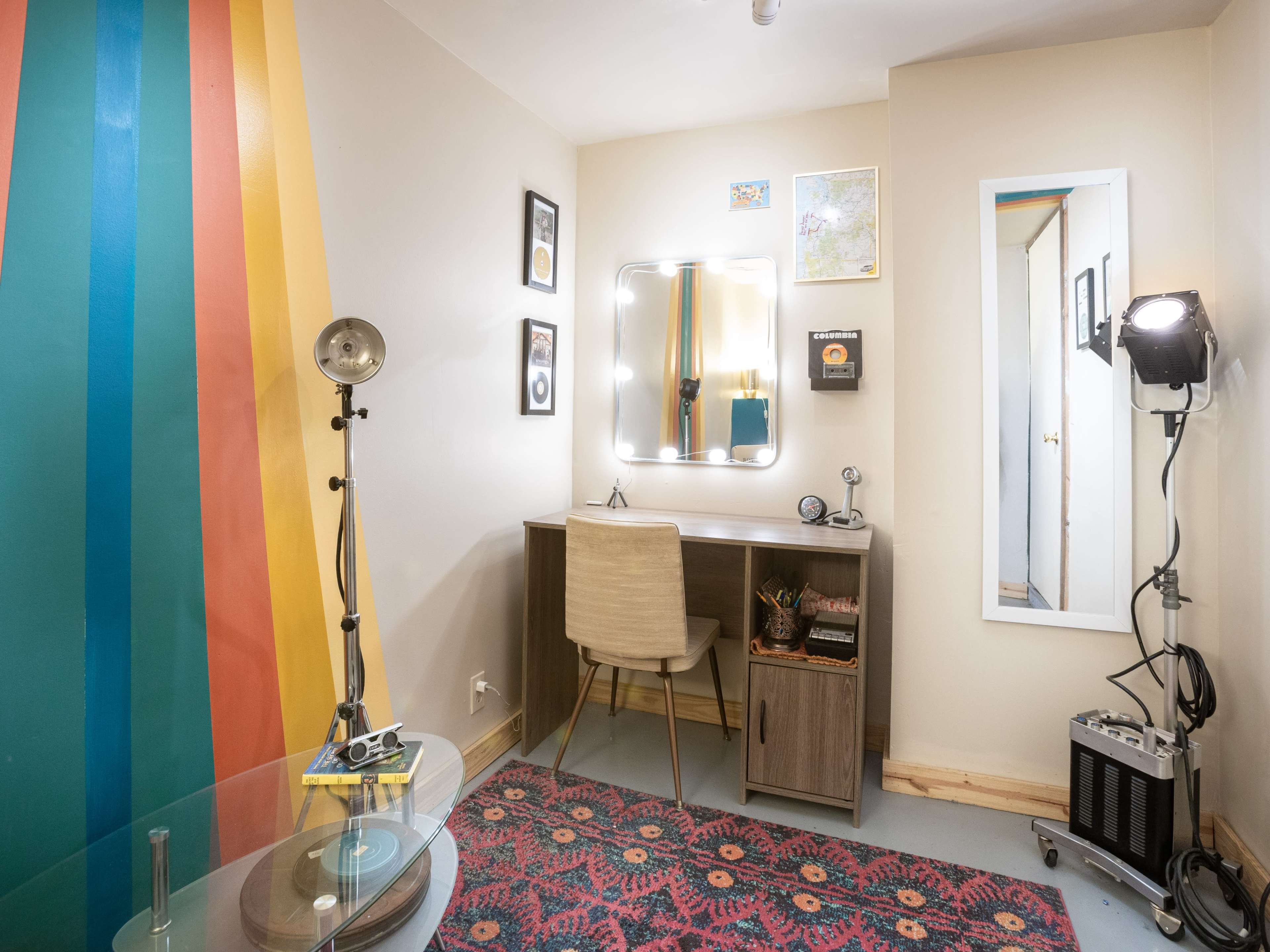 A small room featuring a desk with a mirror, a chair, a record player on a glass table, colorful striped walls, and a light fixture.