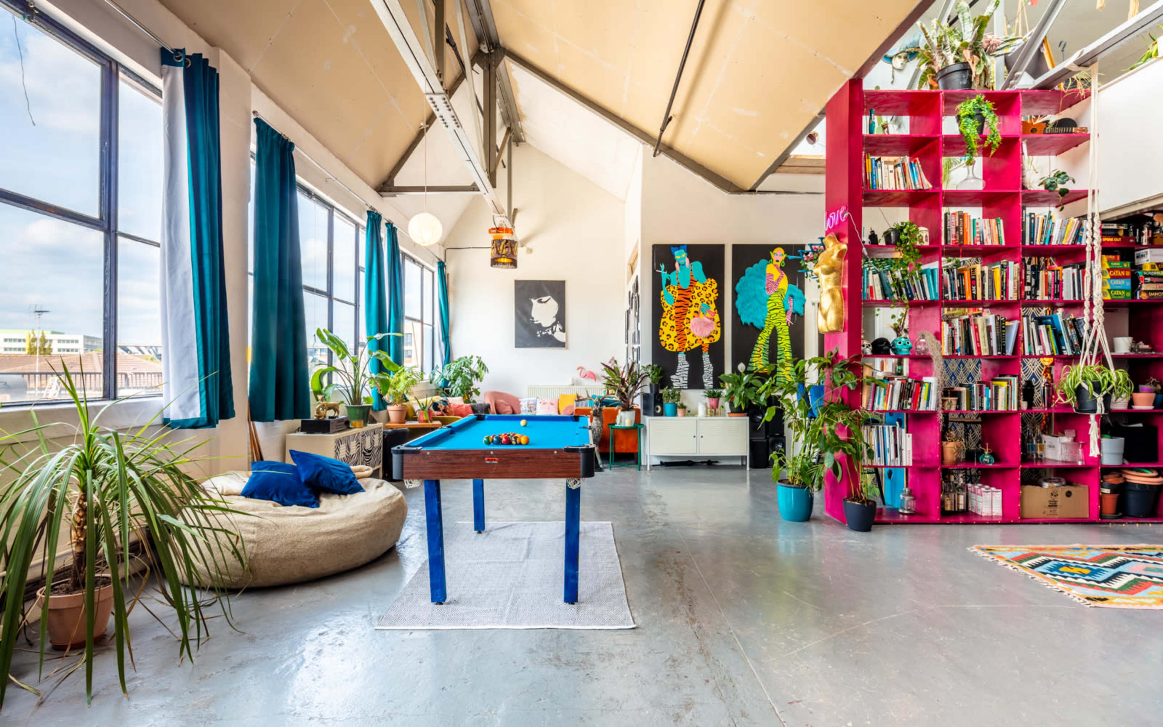 A spacious, bright room featuring a blue pool table, colorful bookshelves filled with books, and various indoor plants.
