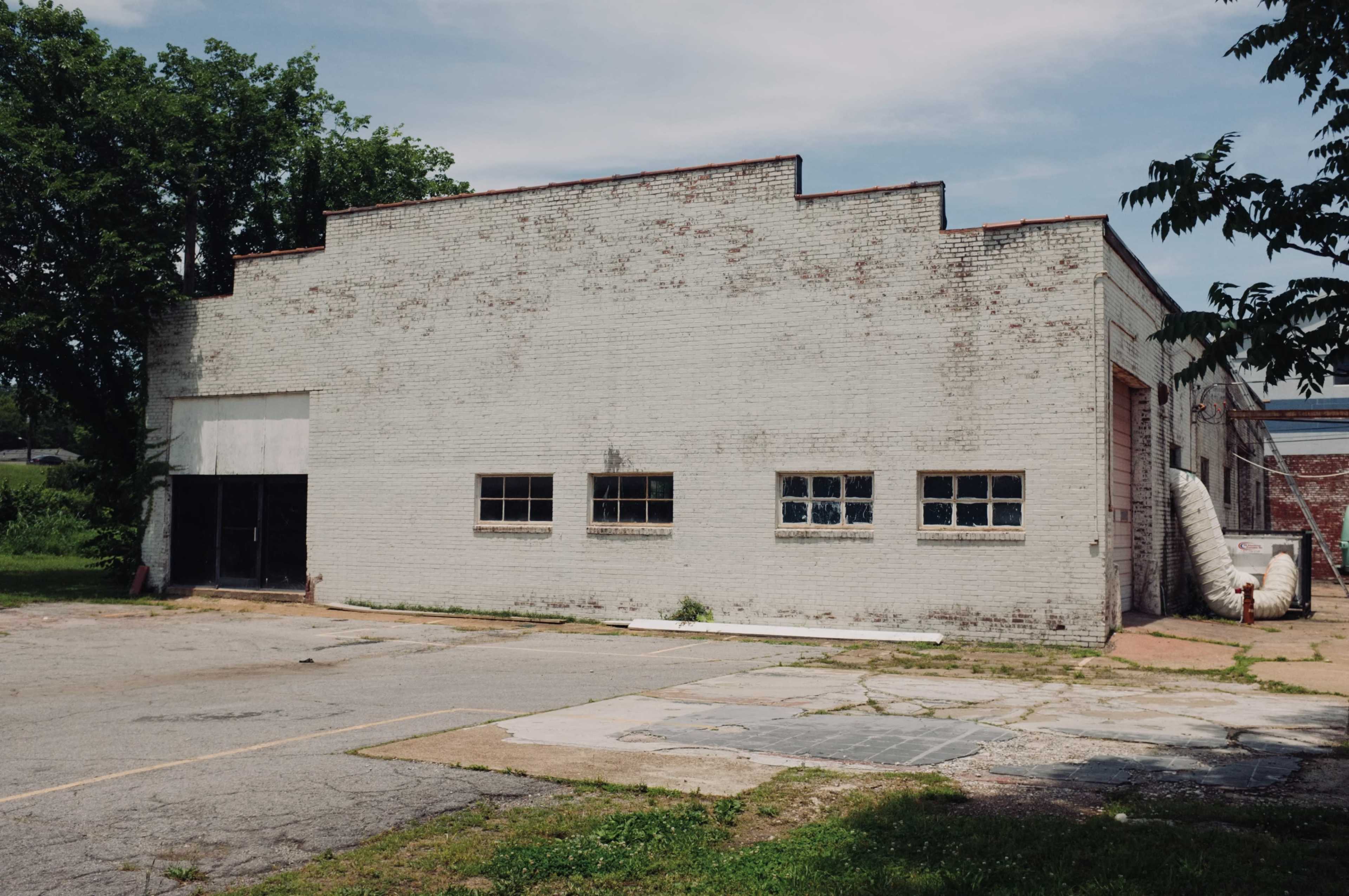 The image shows a weathered, white brick building with several windows and an adjacent empty parking lot surrounded by overgrown grass.