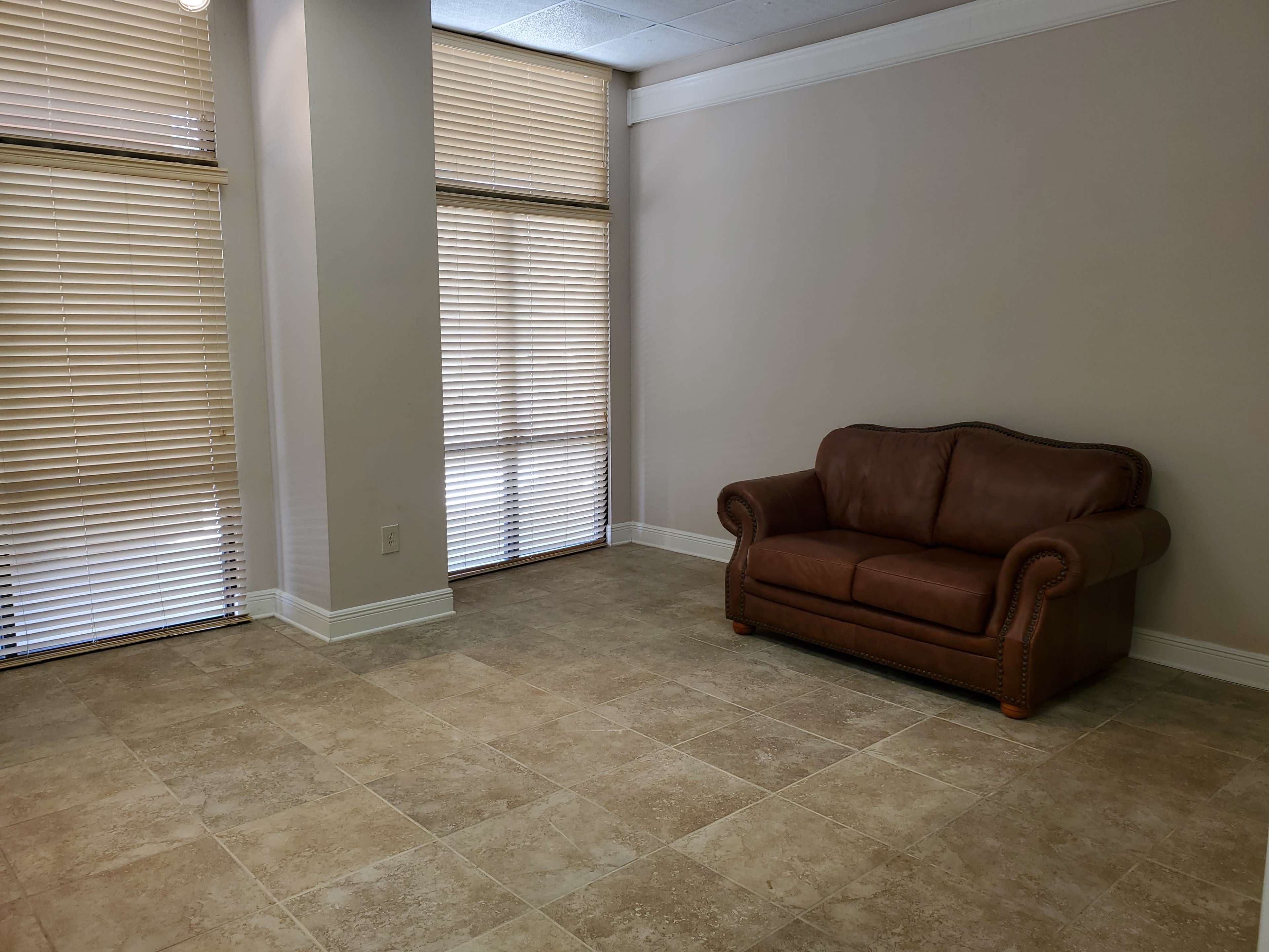 The image shows a brown leather couch placed against a wall in a room with tiled flooring and two windows covered by blinds.