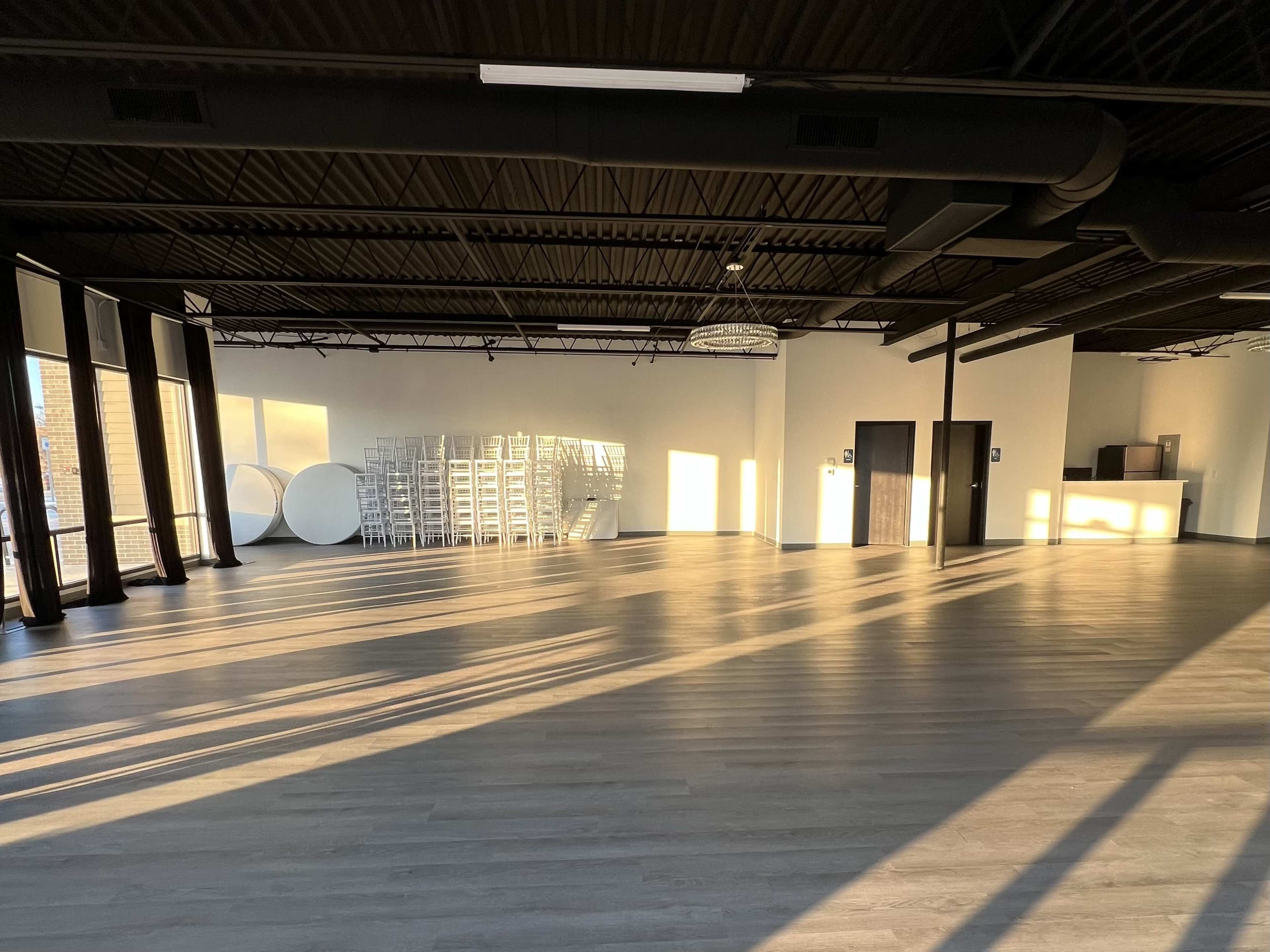 The image shows a spacious, well-lit interior with wooden flooring, large windows casting shadows, and a stack of white chairs in the background.