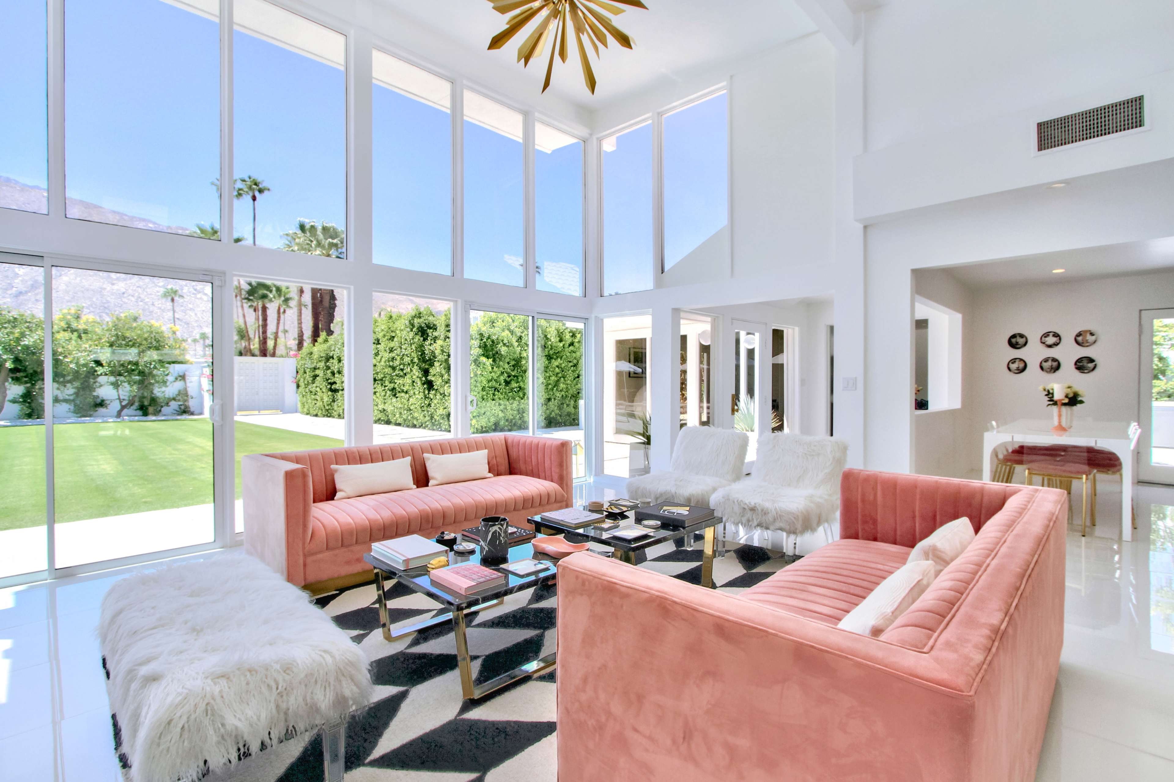 A bright living room featuring two pink couches, a black and white rug, and large windows overlooking a green yard.