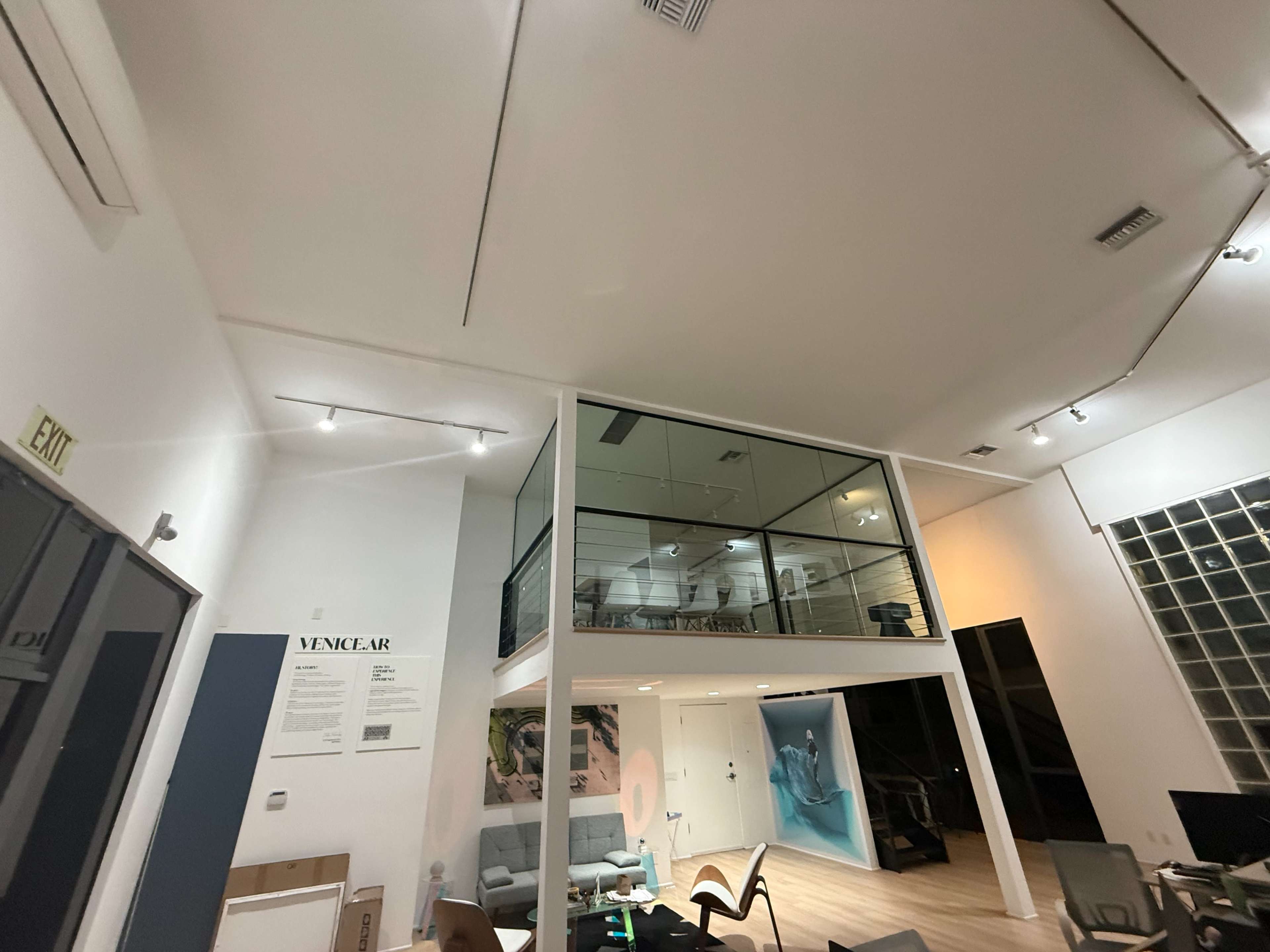 The image shows a spacious office interior with a glass-walled upper level, modern furniture, and a mix of white and light-colored walls.