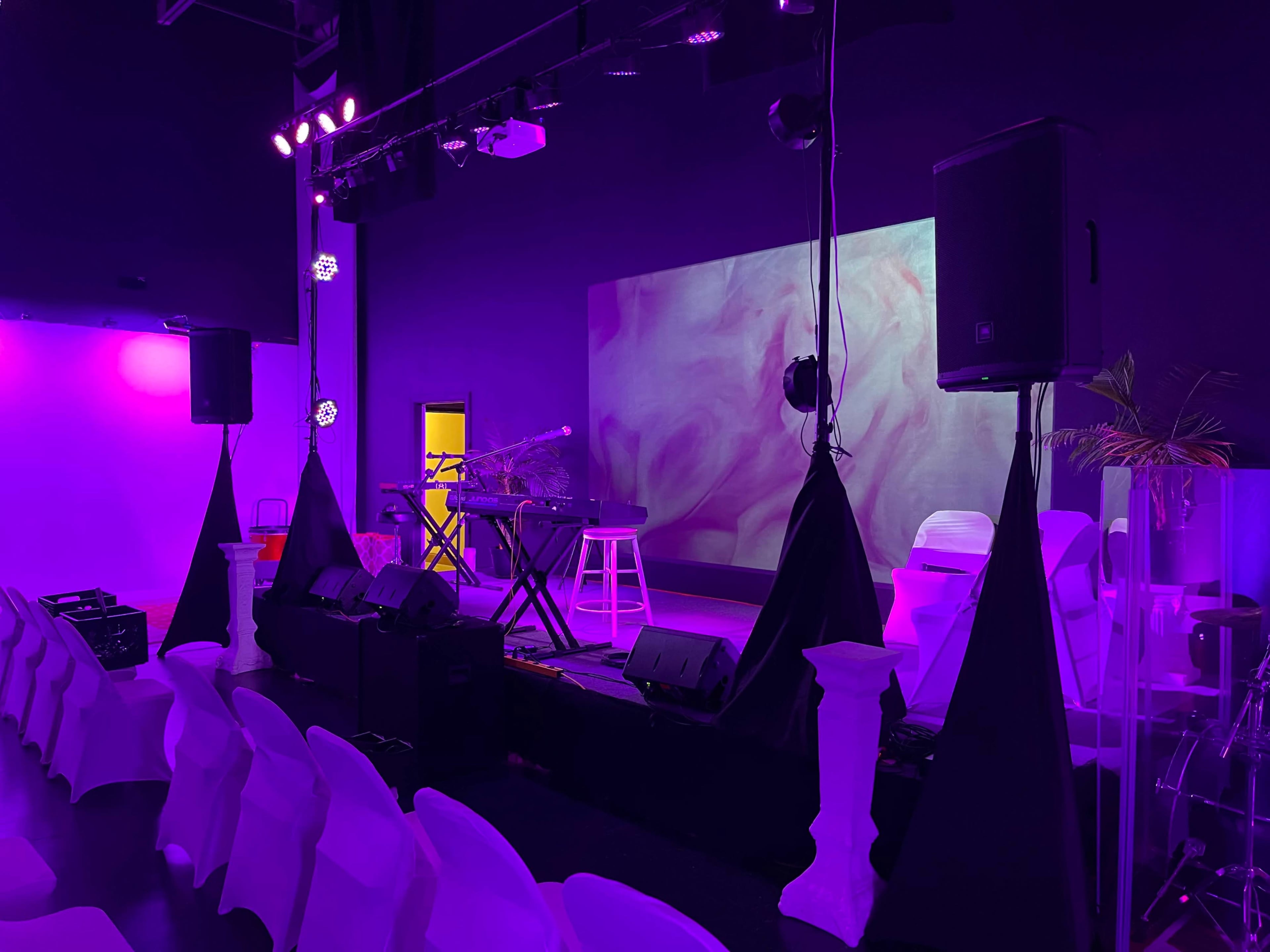 A stage setup in a dimly lit venue with purple lighting, featuring a keyboard, a projector screen with abstract visuals, and rows of white chairs facing the stage.
