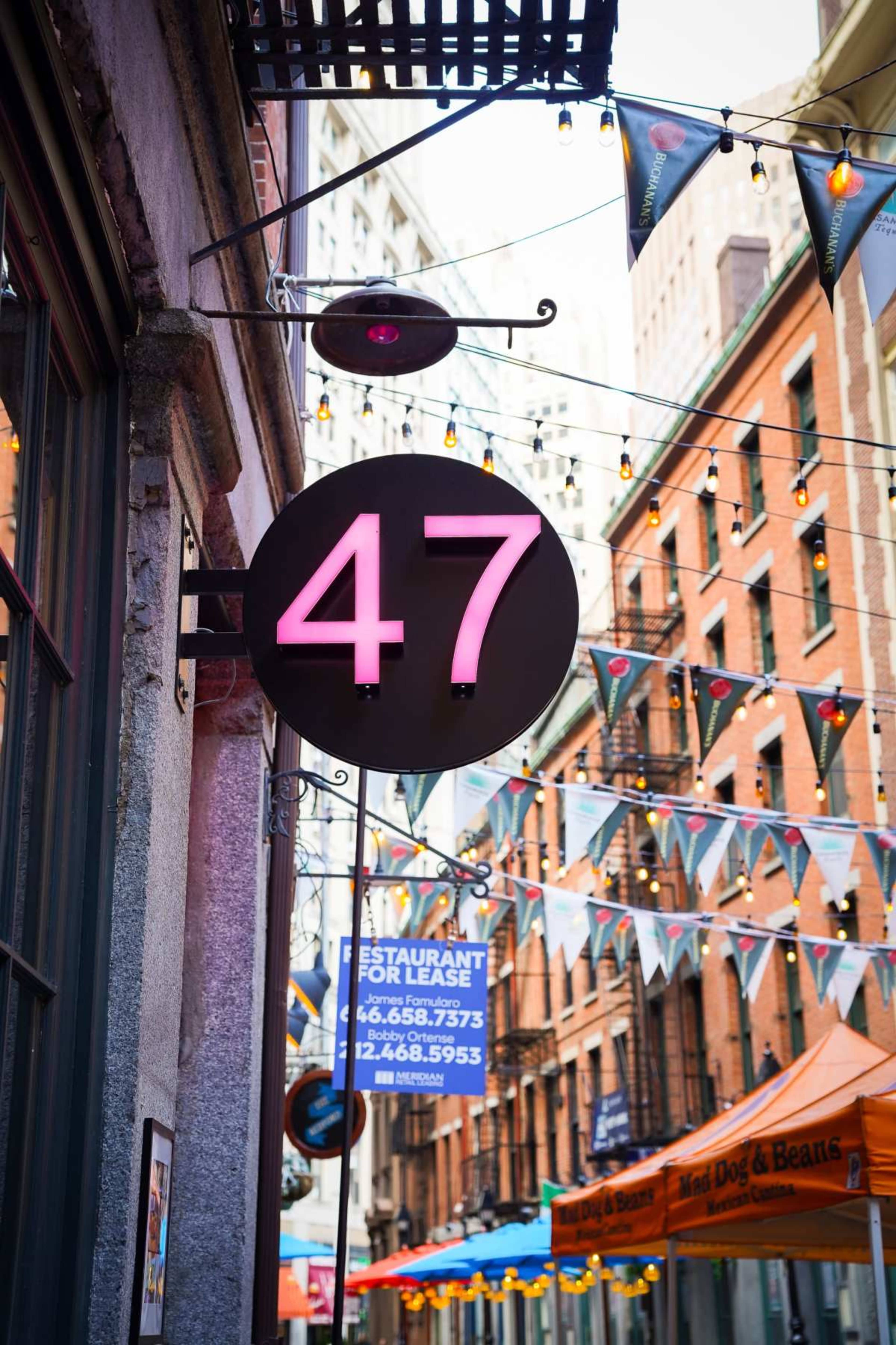 A large circular sign with the number "47" is displayed outside a building in a colorful alleyway decorated with string lights and festive bunting.