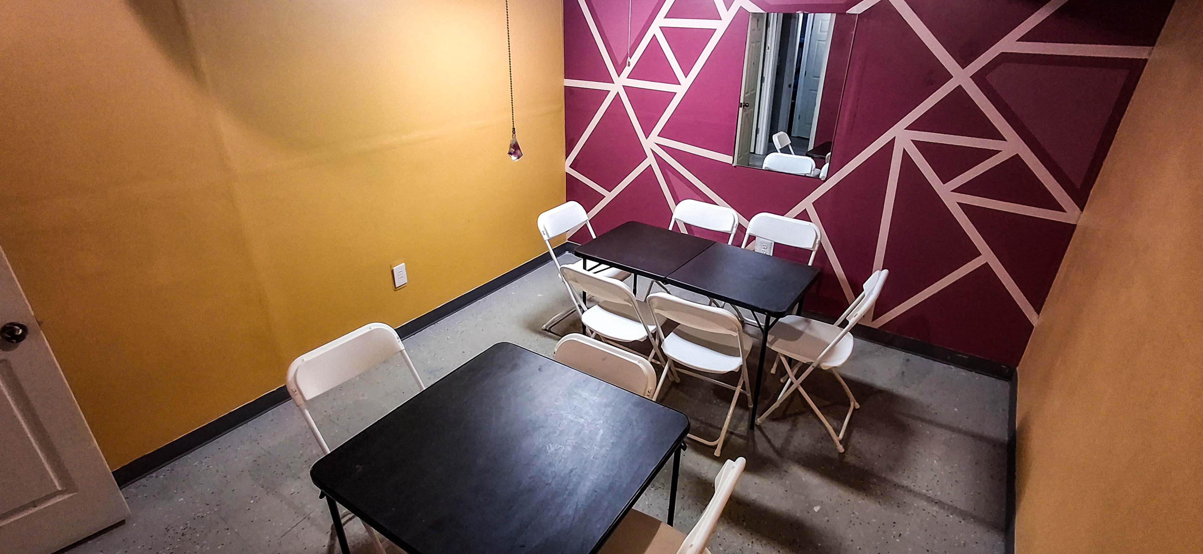 The image shows a small meeting room with two black tables and several white folding chairs arranged around them, featuring a colorful geometric patterned wall and a mirror.