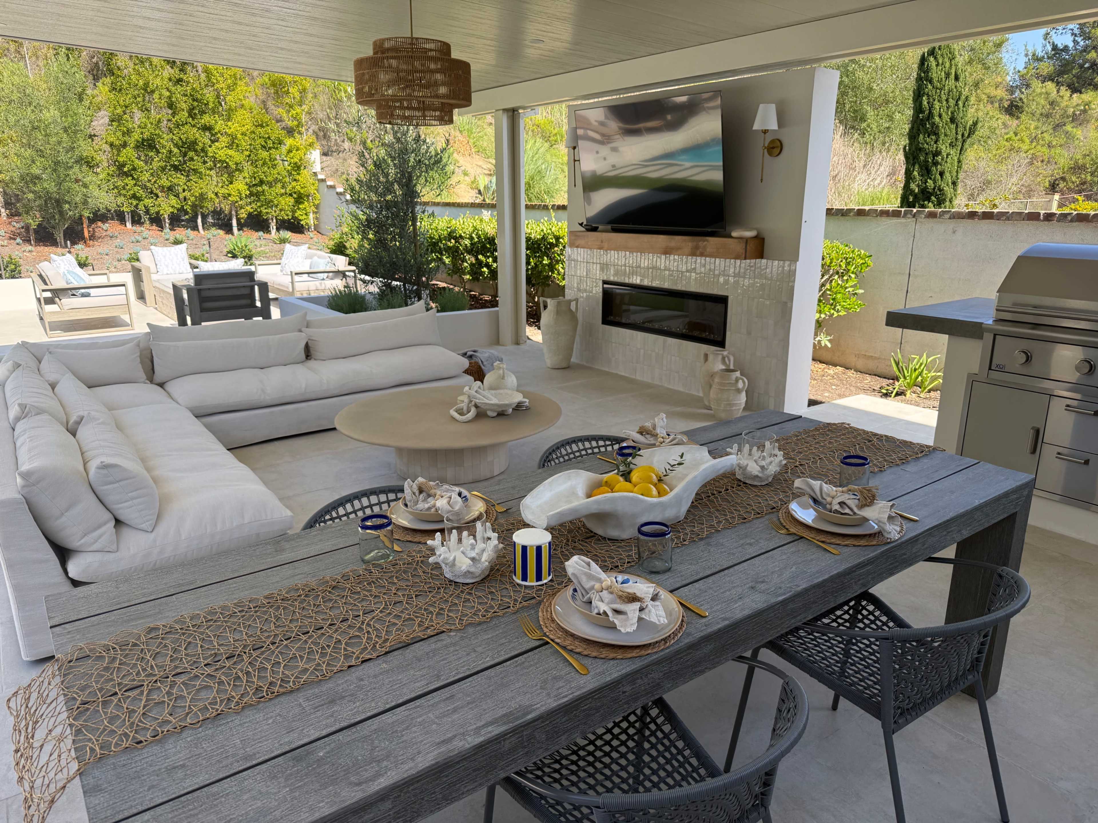 The image shows a modern outdoor patio area featuring a large sectional sofa, a coffee table, a dining table set with dishes, and a built-in grill, all under a covered roof.