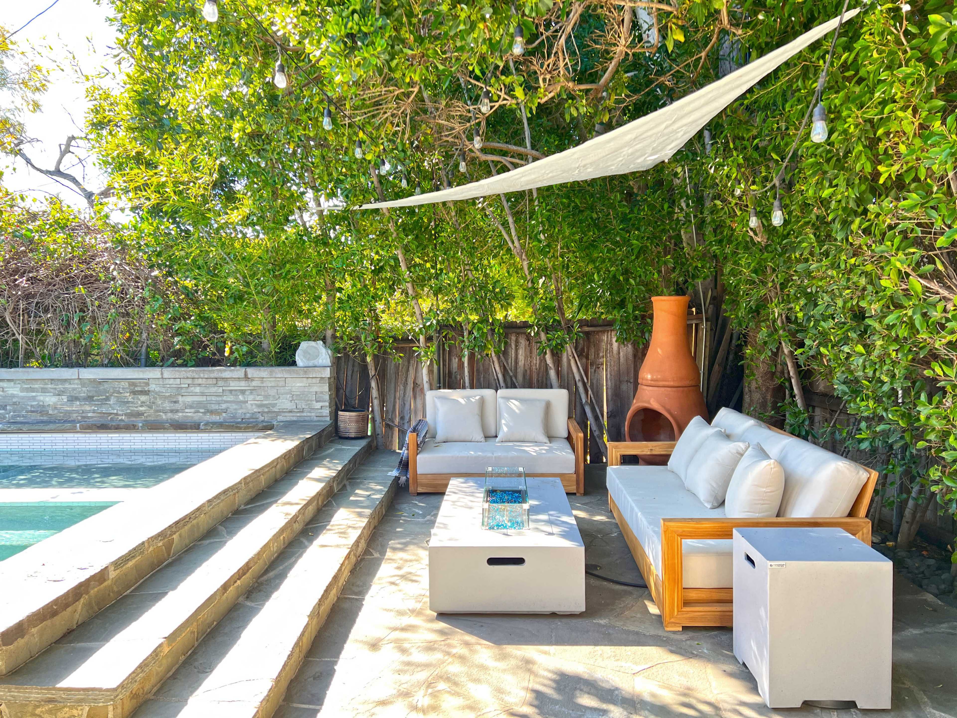 A cozy outdoor seating area with a sofa, coffee table, and a terracotta chiminea beside a swimming pool, all shaded by a canopy and surrounded by lush greenery.