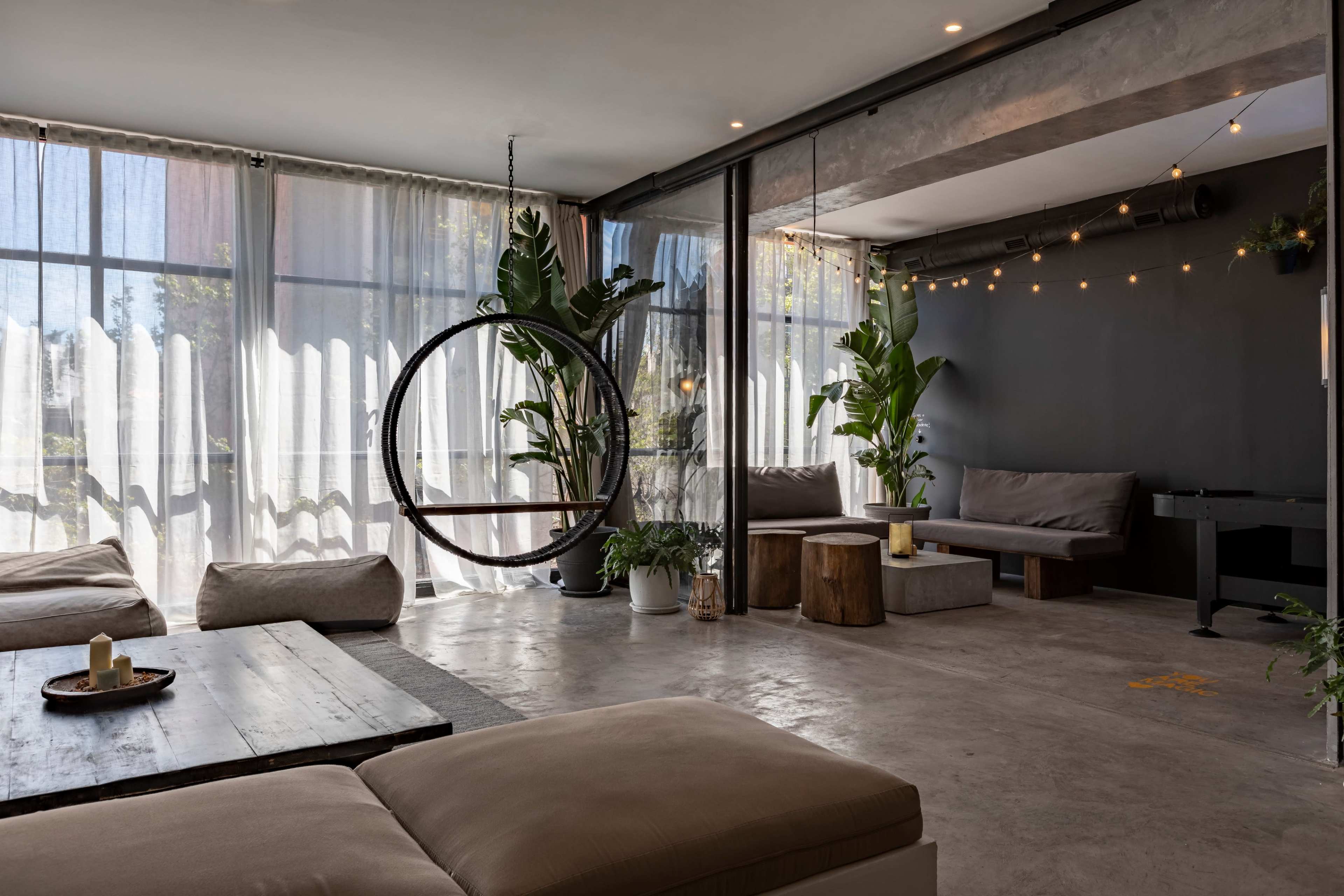 A spacious, modern lounge area features a circular hanging chair, potted plants, and comfortable seating with large windows allowing natural light to fill the space.