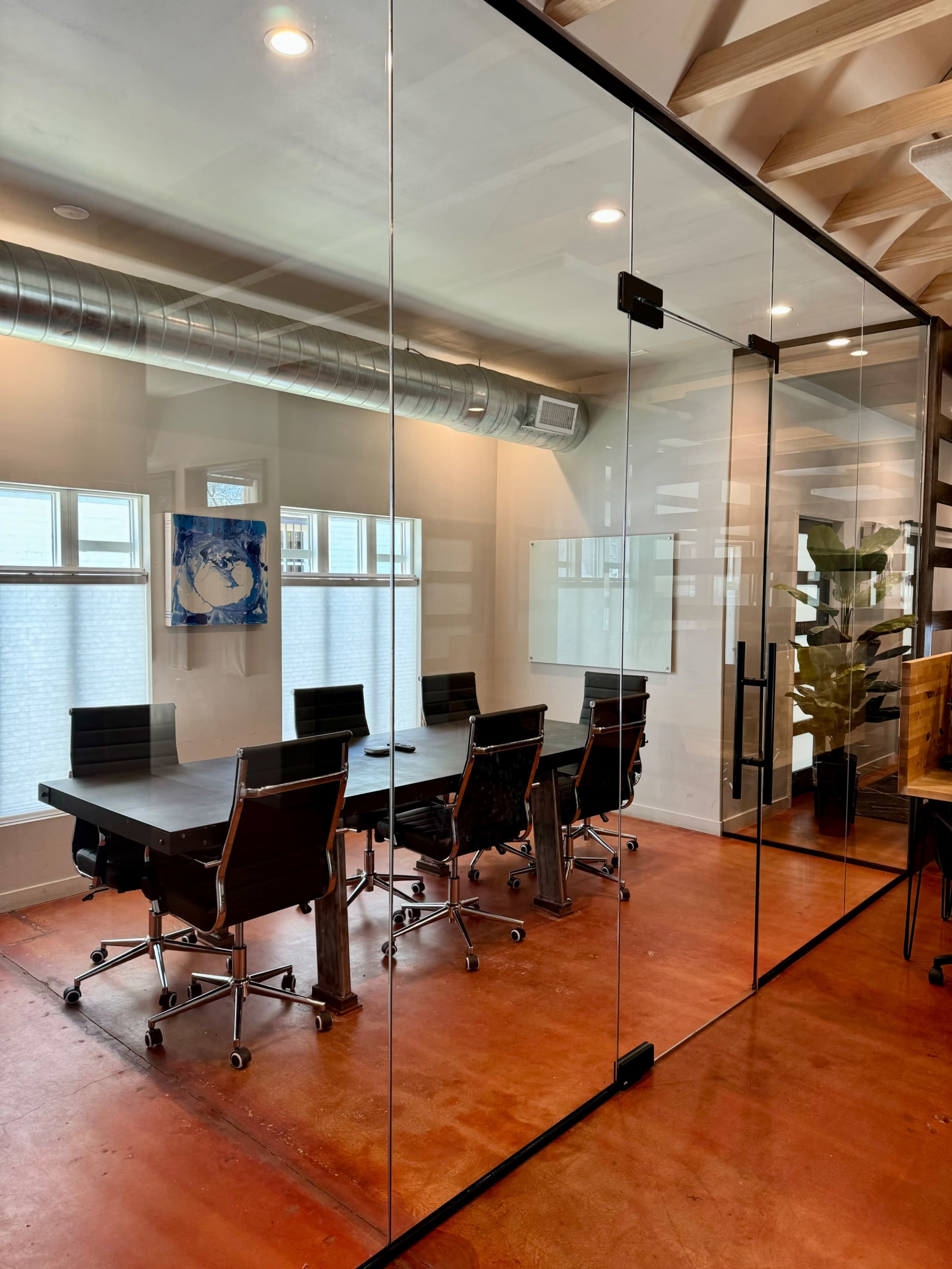 The image shows a modern conference room with a large glass wall, featuring a long table surrounded by black rolling chairs and a decorative plant in the corner.