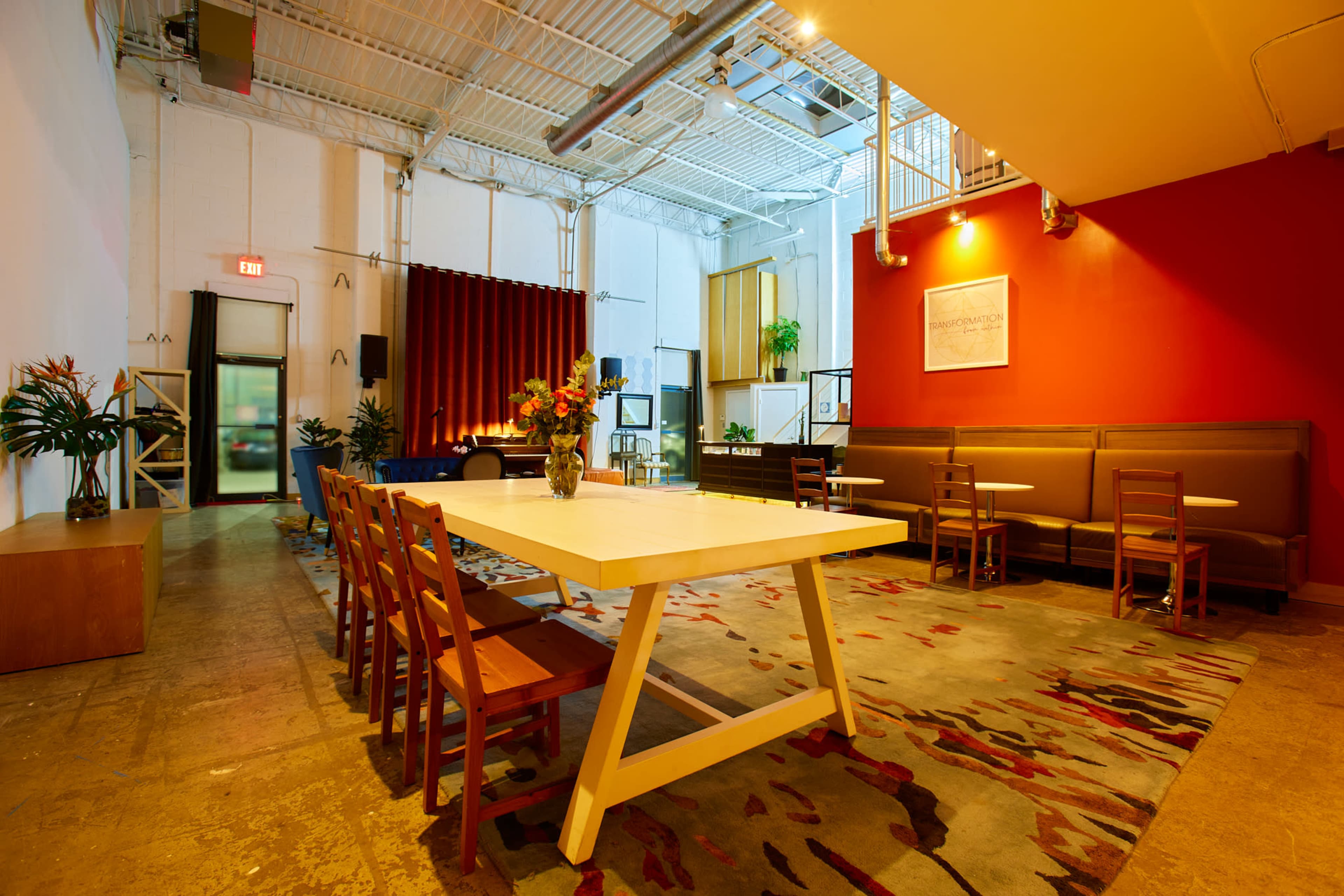 A well-lit, contemporary gathering space with a large table, several chairs, potted plants, and a stage area in the background featuring a red wall.