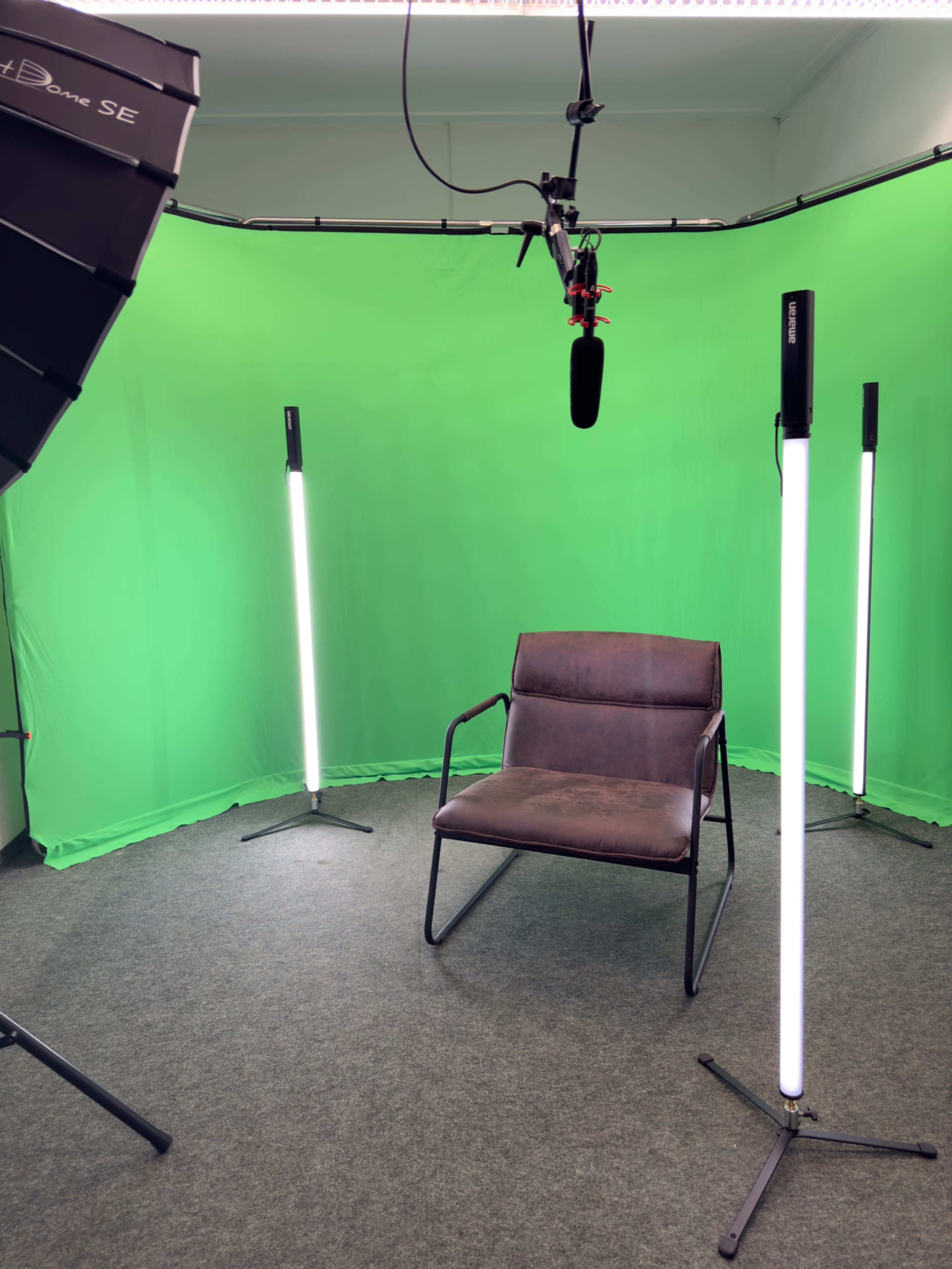 The image shows a preparation area for video recording, featuring a green backdrop, a single chair in the center, and illumination from tall LED lights.