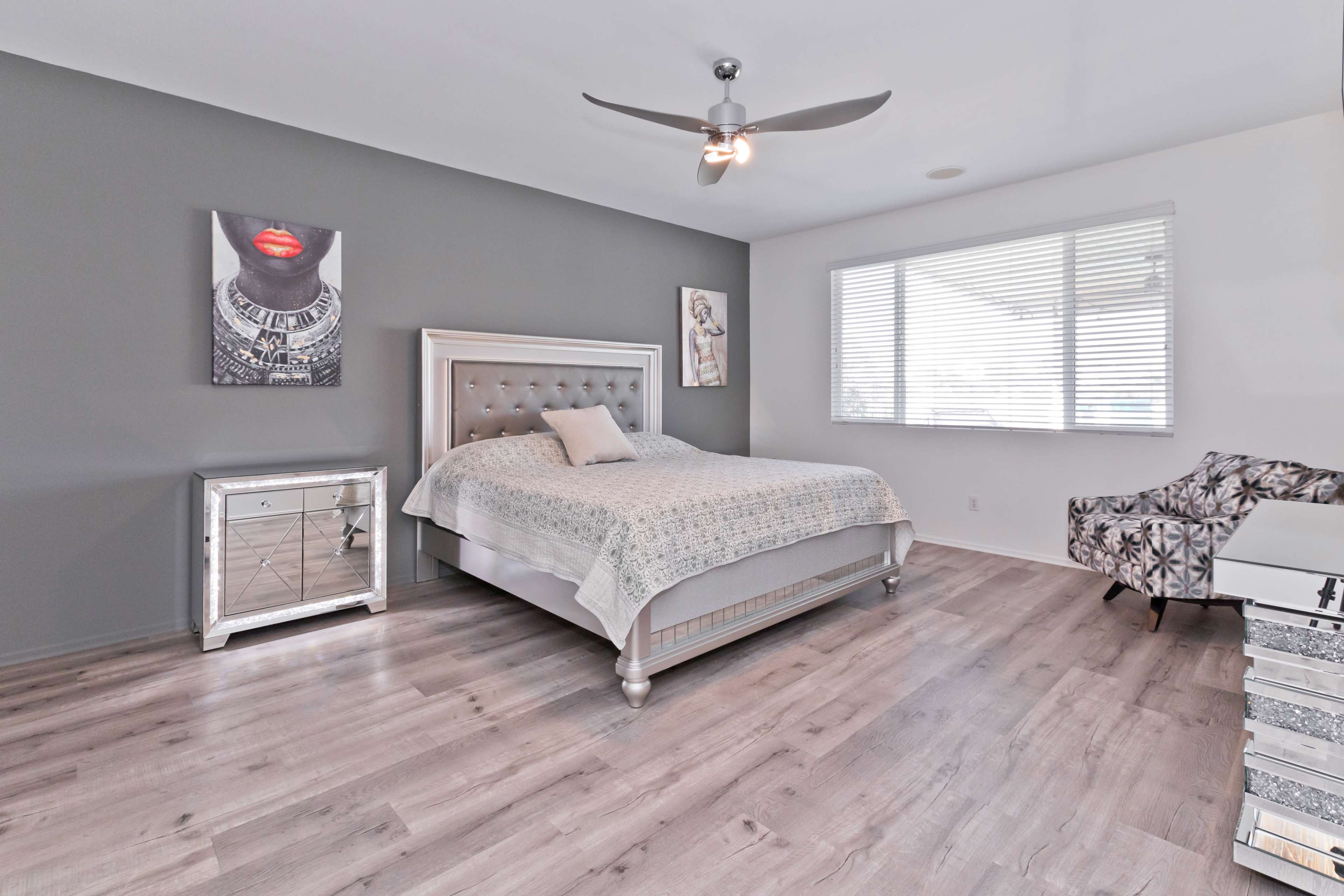 A spacious bedroom featuring a bed with a light-colored quilt, a mirrored nightstand, a patterned chair, and large windows with white blinds.