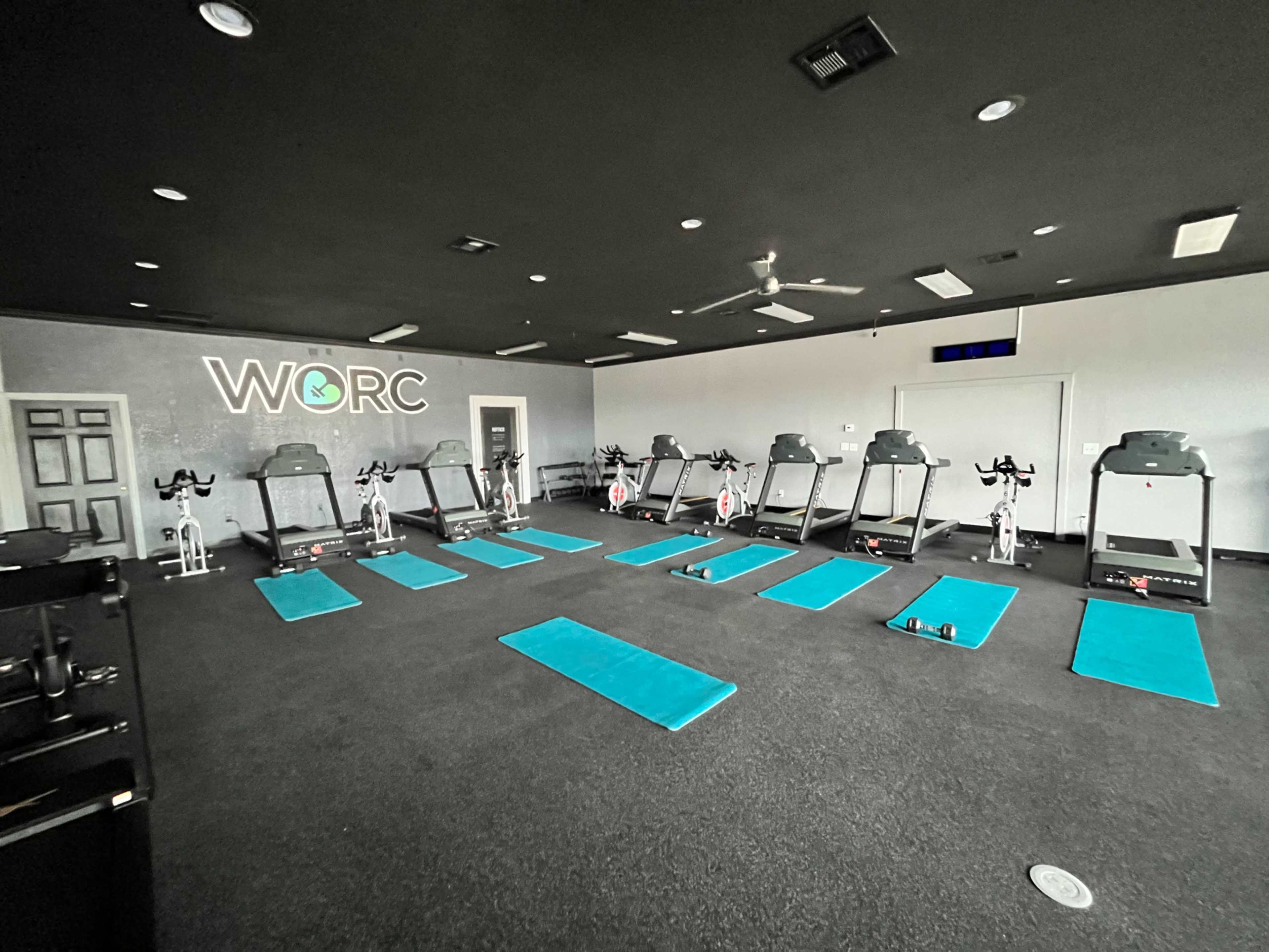 The image shows an indoor fitness studio equipped with several exercise bikes, workout mats, and a branding wall labeled "WORC."