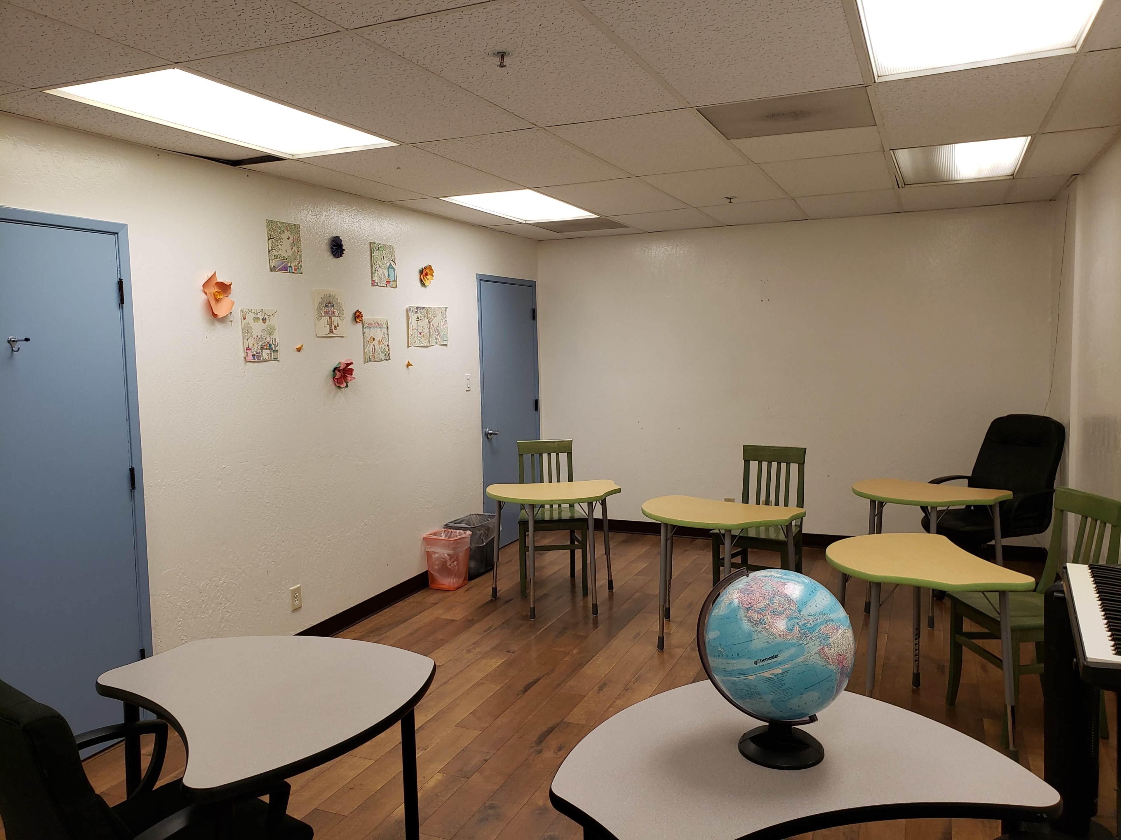 A small classroom with several tables and chairs arranged around a globe and a keyboard, along with artwork displayed on the wall.
