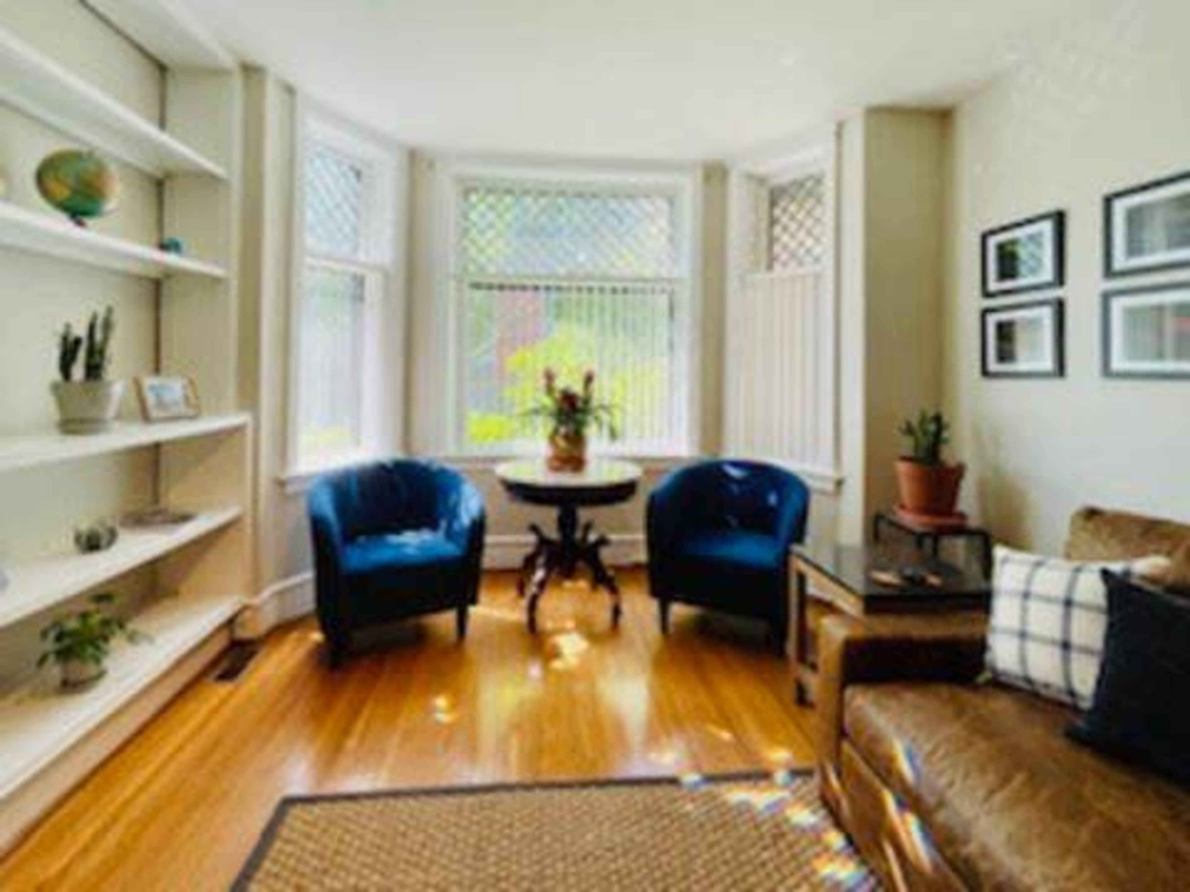 A cozy living room features two blue chairs, a small round table, and a couch, illuminated by natural light from bay windows.