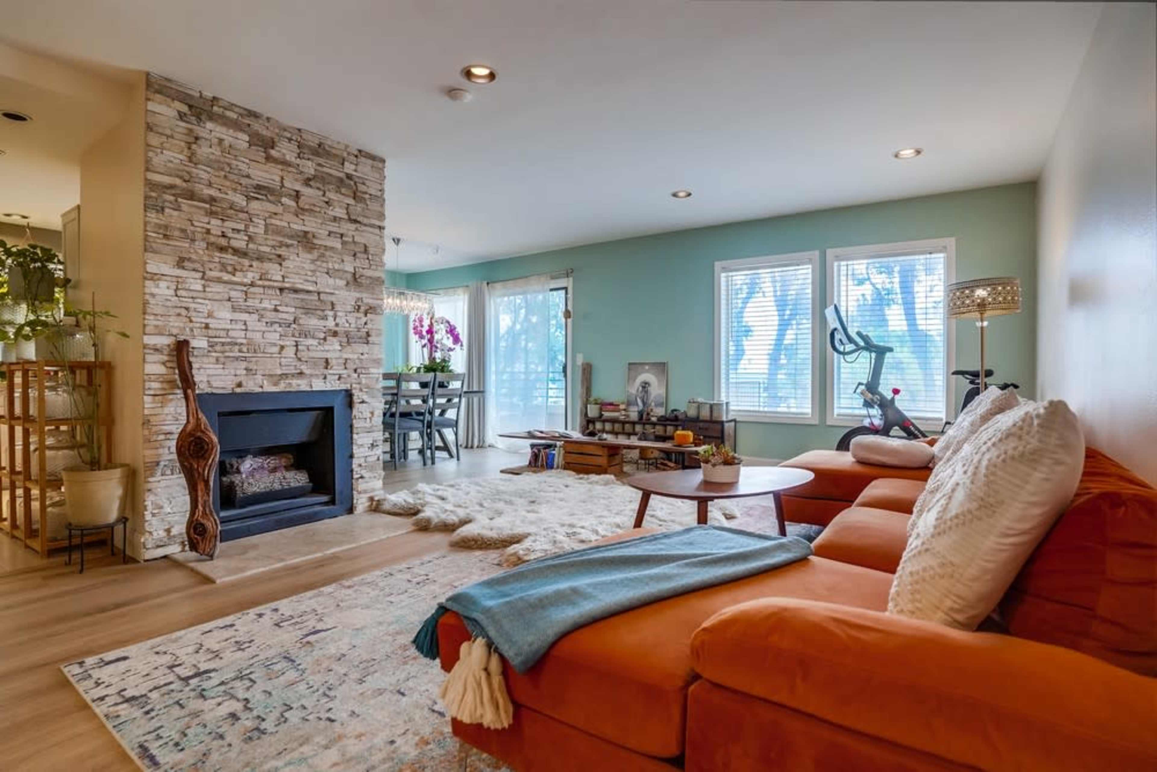 A cozy living room featuring an orange sectional couch, a stone fireplace, and large windows with a view of greenery outside.