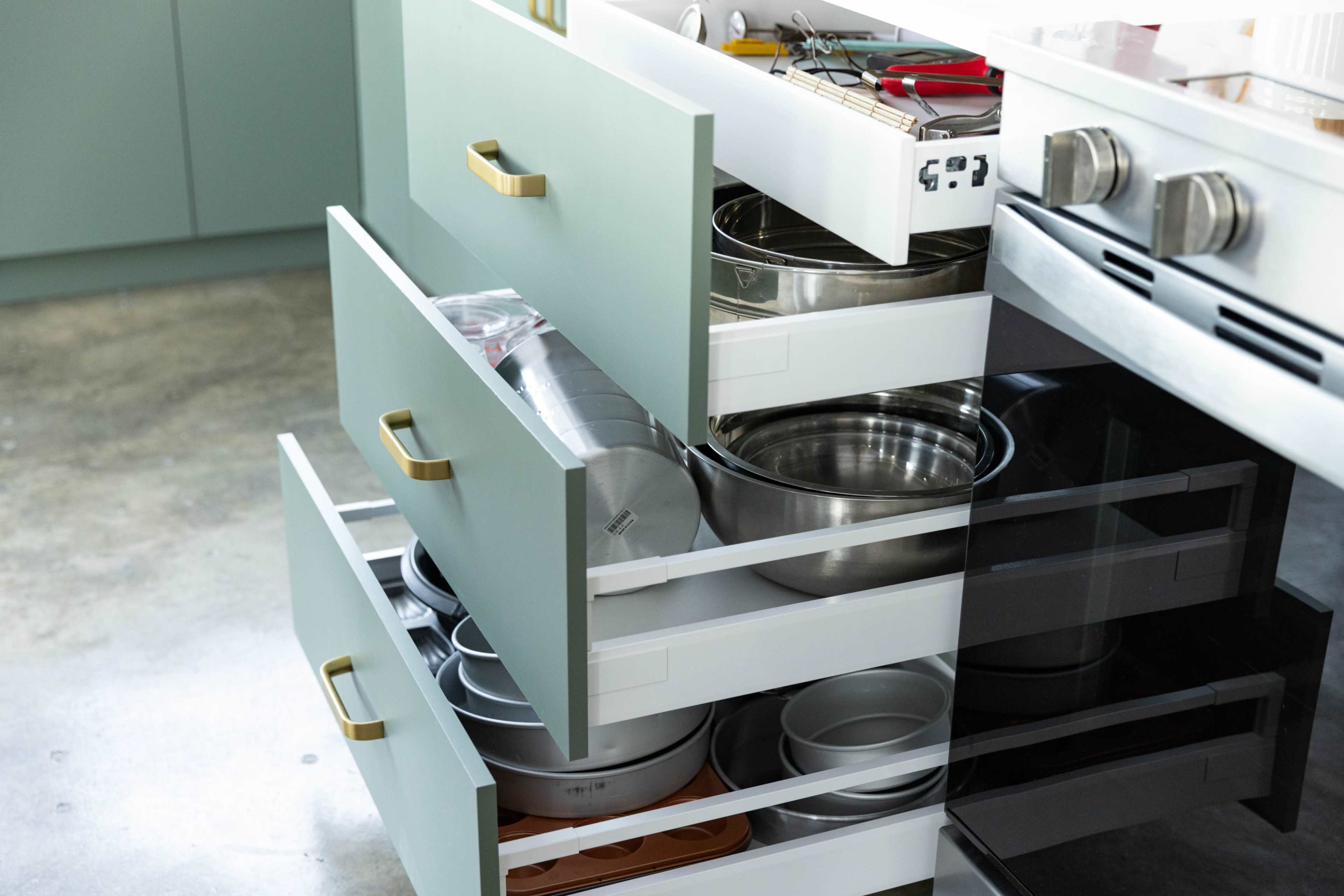 A kitchen cabinet with three open drawers contains various metal bowls, baking pans, and utensils.