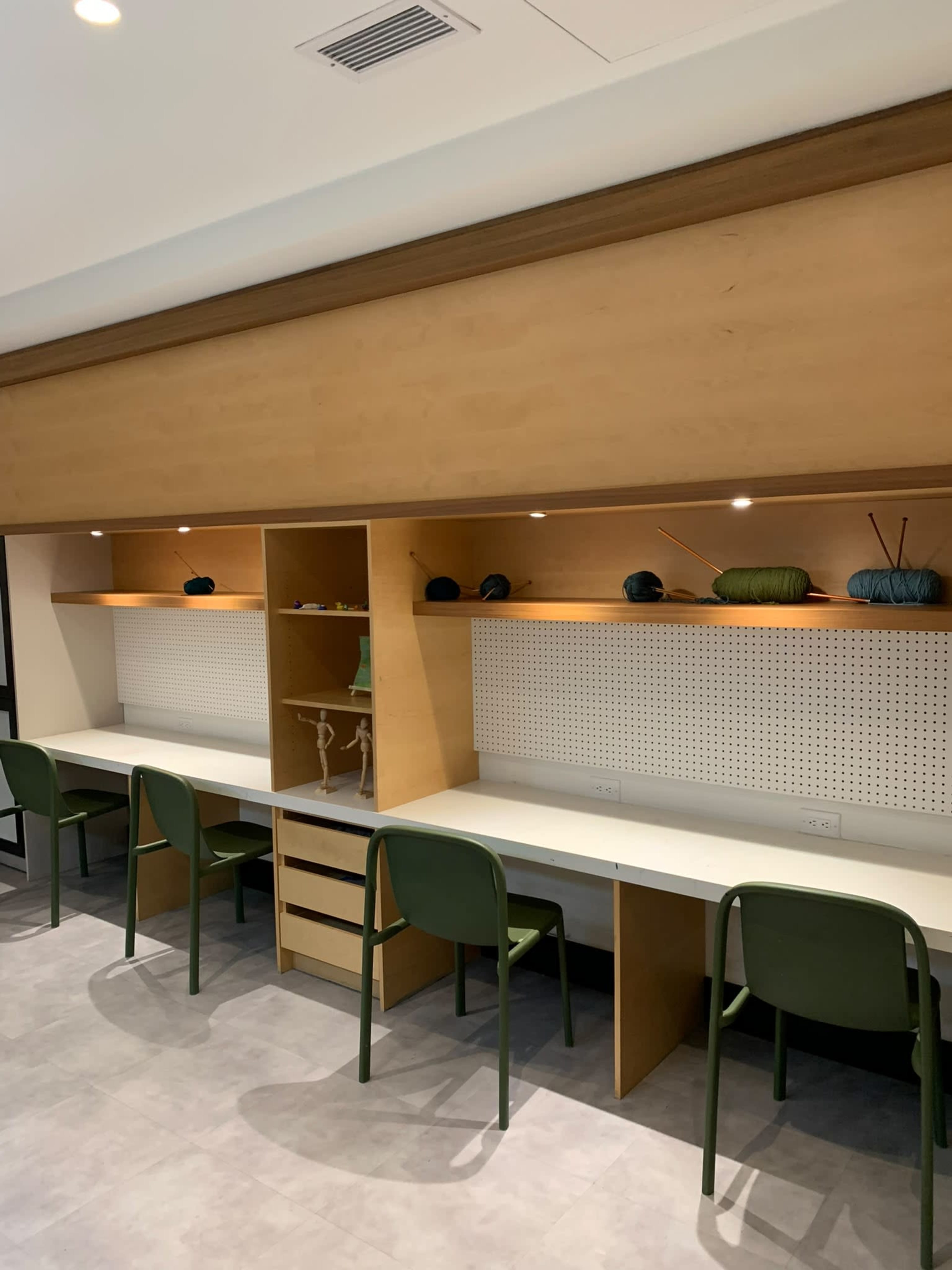 The image shows a clean, minimalist room featuring wooden shelves, a long white table with several green chairs, and some decorative items on the shelves.