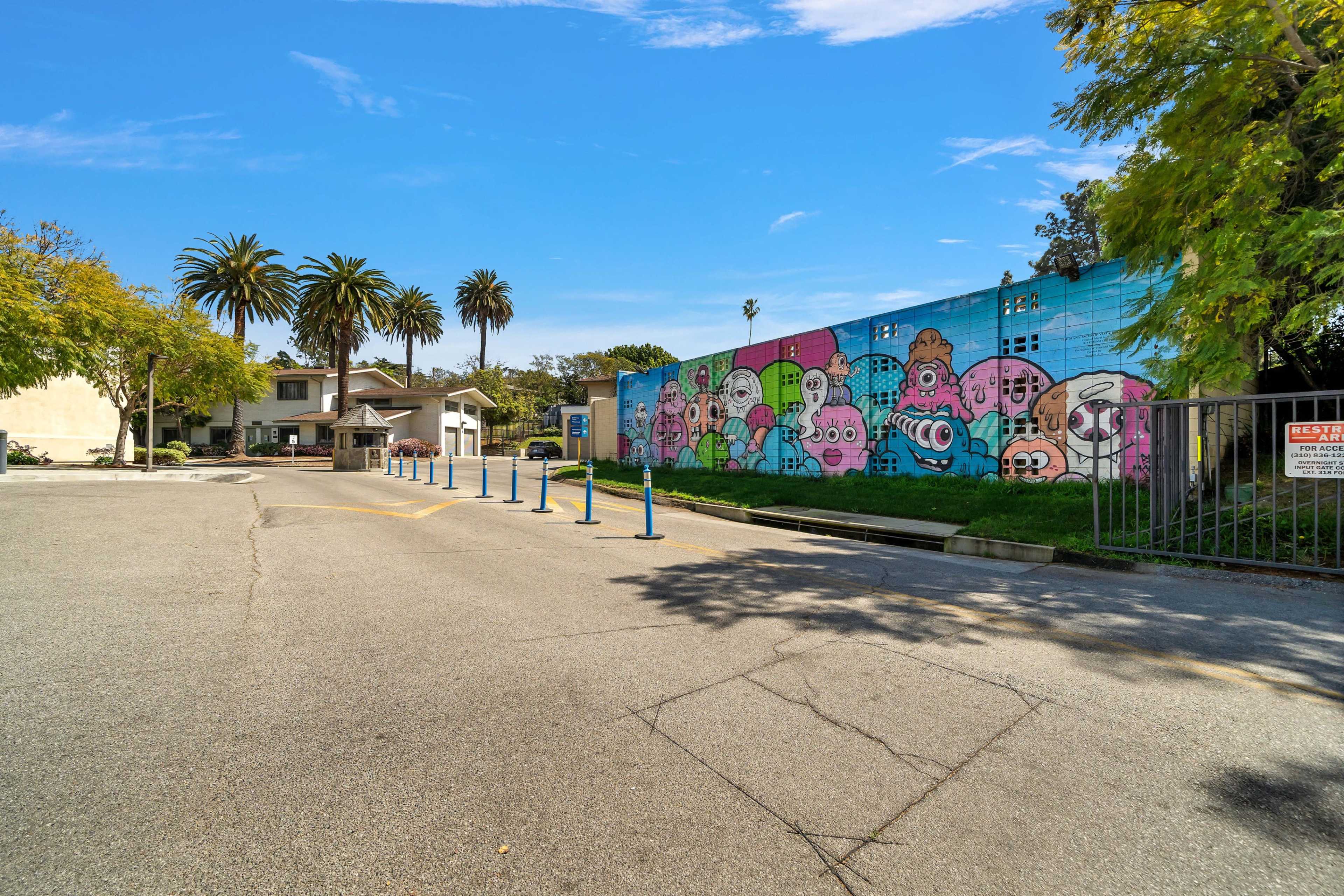 A street with a colorful mural on a wall, lined by palm trees and parking poles.