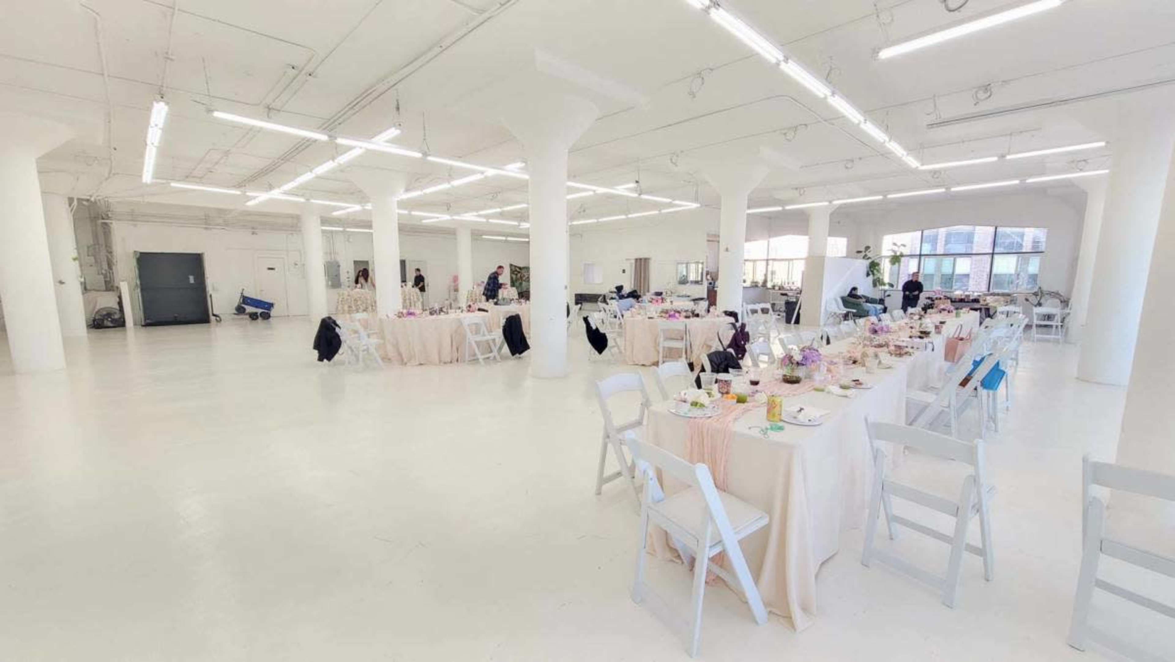 The image shows a spacious indoor venue with several round tables set for a meal, adorned with tablecloths and dishes, and bright overhead lighting.