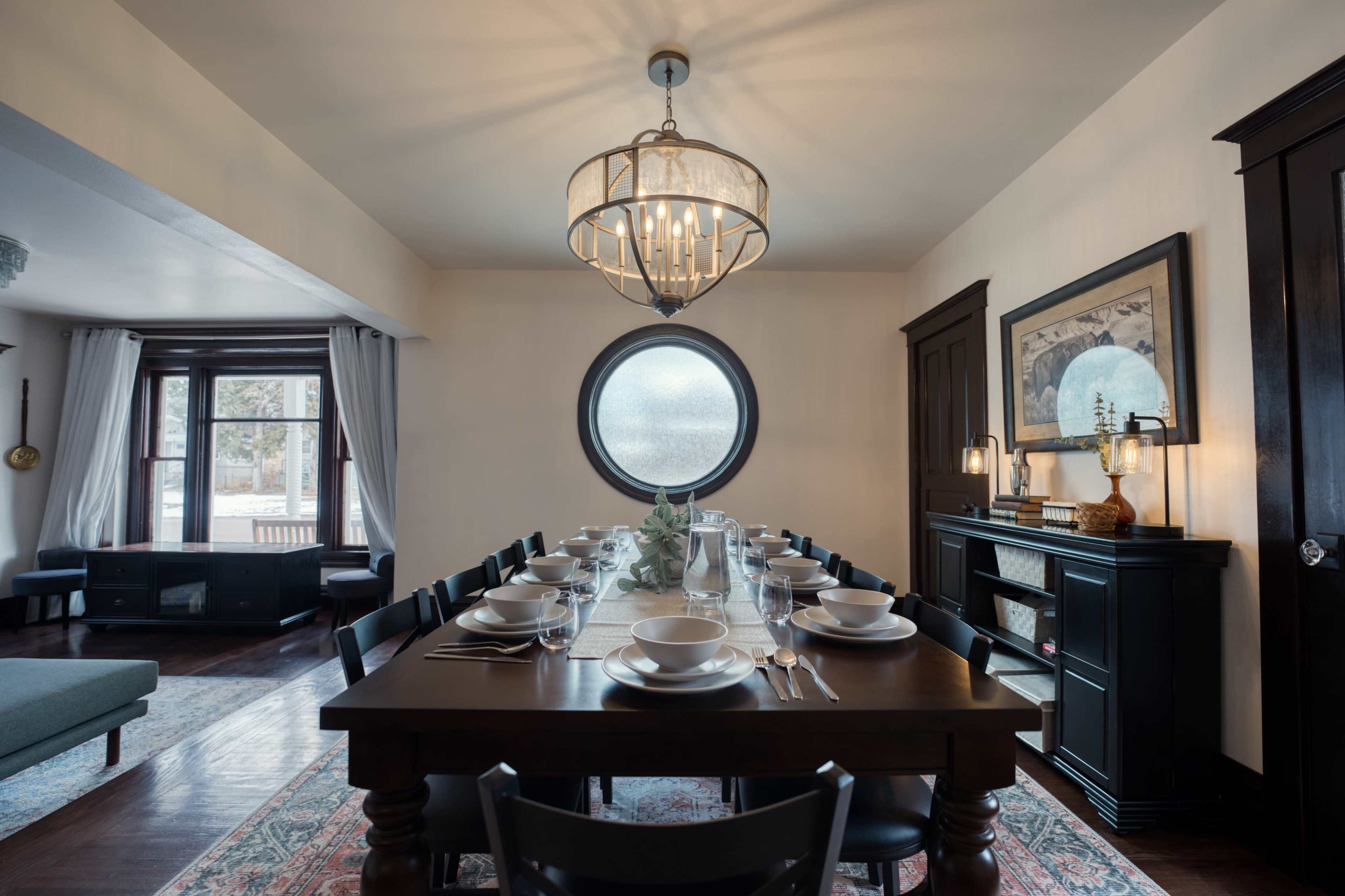 A dining room with a long table set for a meal, featuring a circular window and a chandelier above.