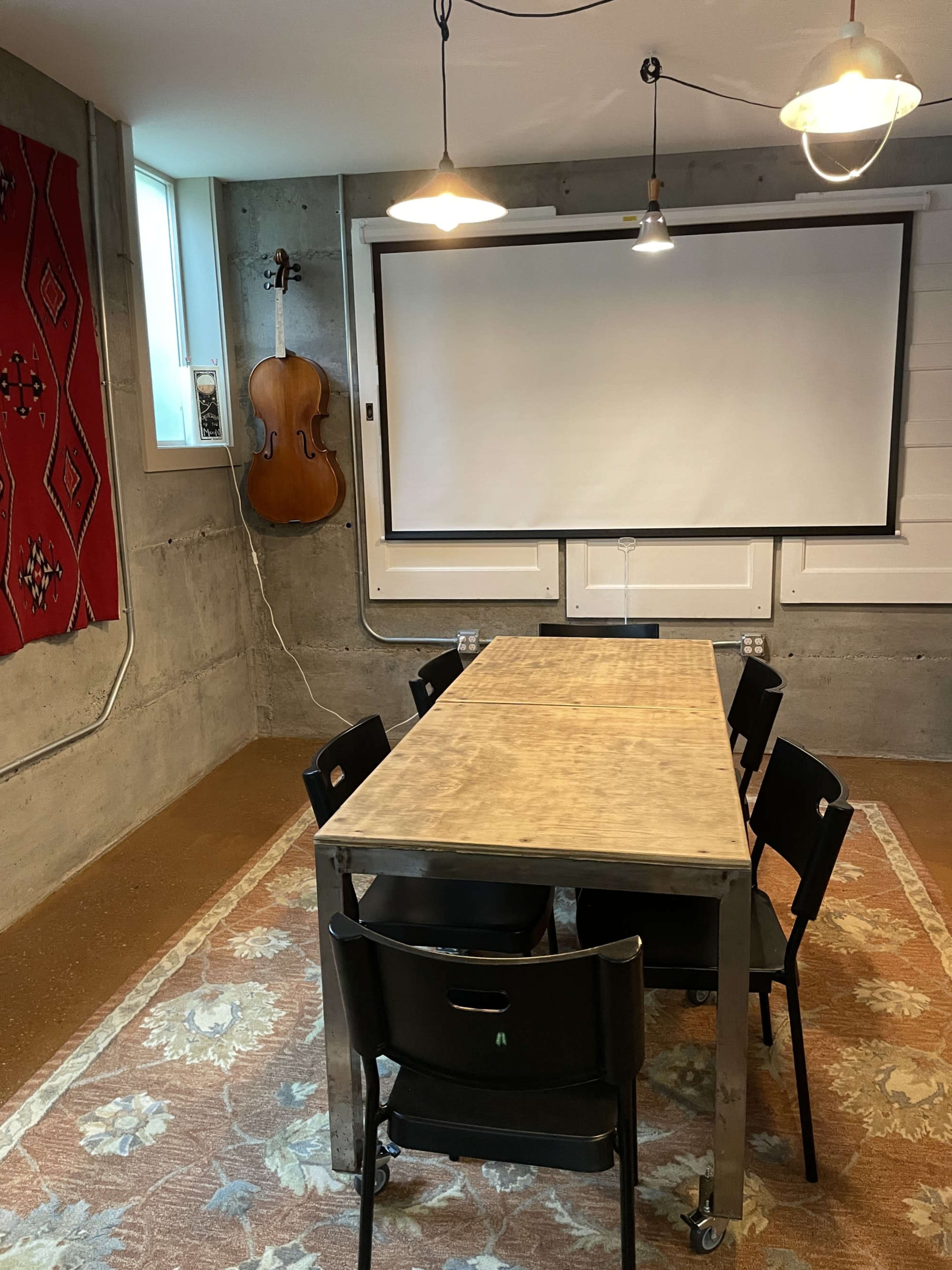 A rectangular table surrounded by black chairs is set in a room with concrete walls, a large screen, and a cello hanging on the wall.