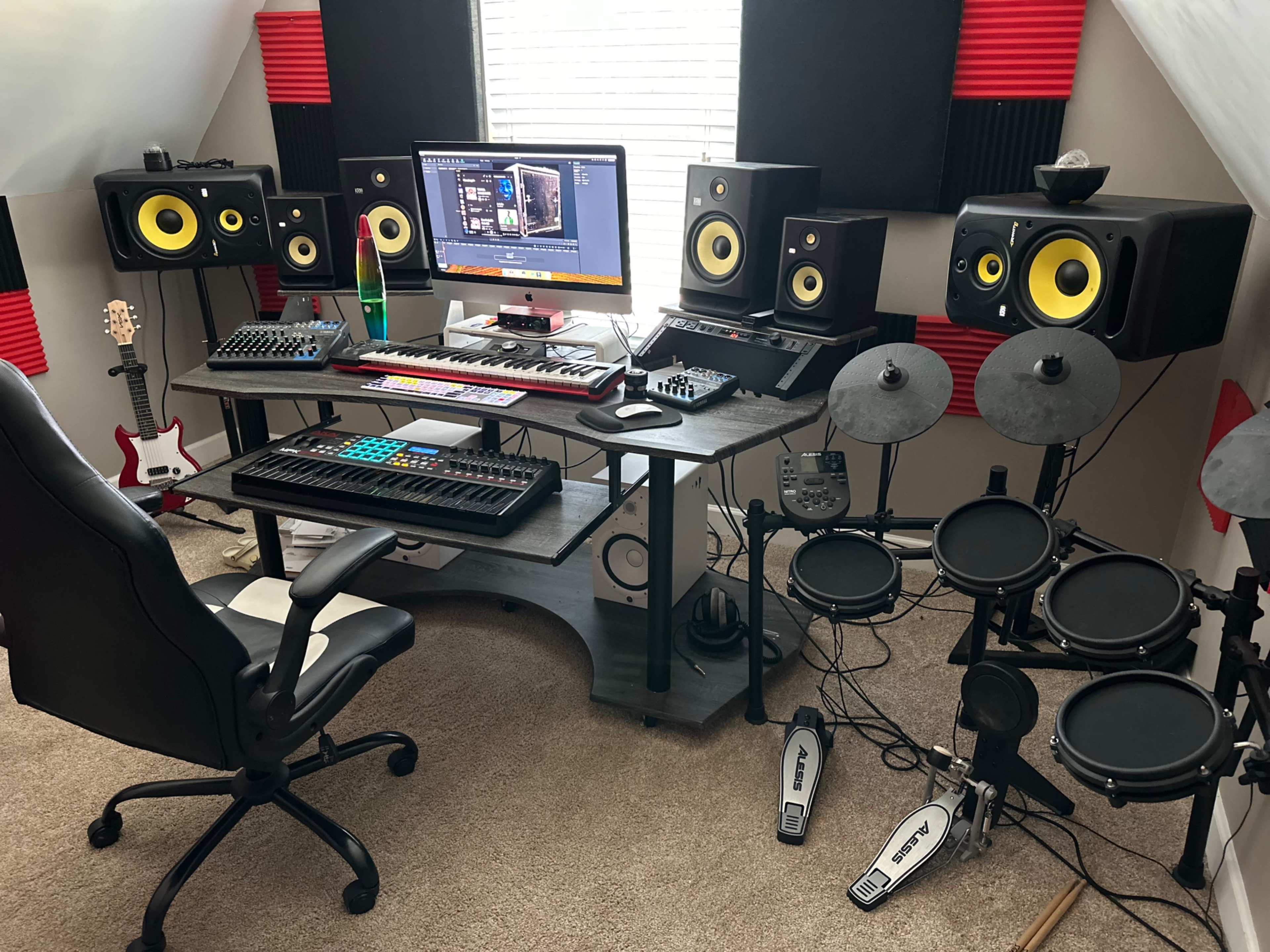 A home music production setup features a computer display, studio monitors, a MIDI keyboard, an audio interface, and electronic drum pads arranged around a desk.