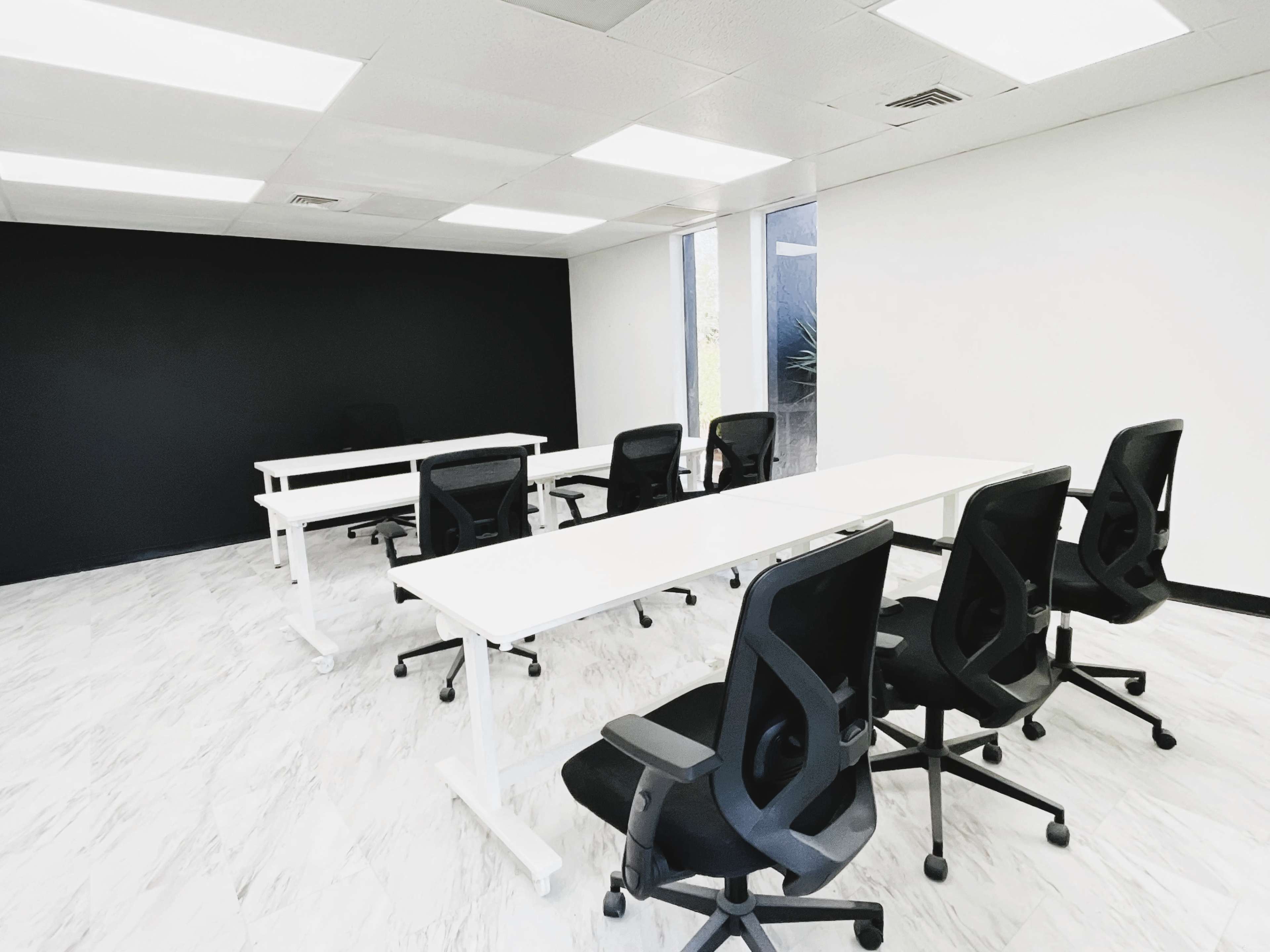 The image shows a modern meeting room with white tables arranged in a U-shape and black ergonomic chairs, featuring a black accent wall and windows letting in natural light.