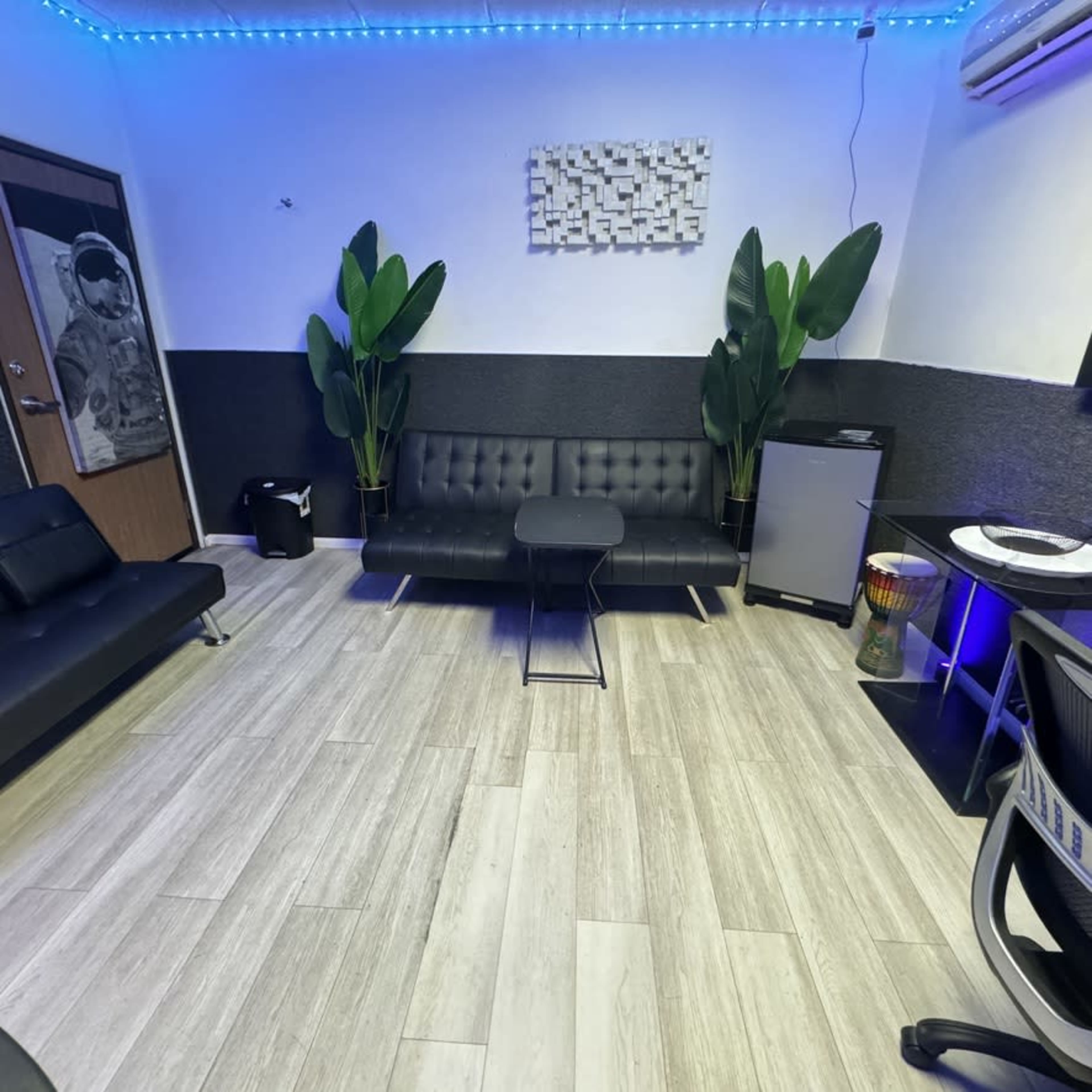 The image shows a modern, minimalist room with a black sofa, a small table, a mini fridge, and potted plants, all under blue LED lighting.
