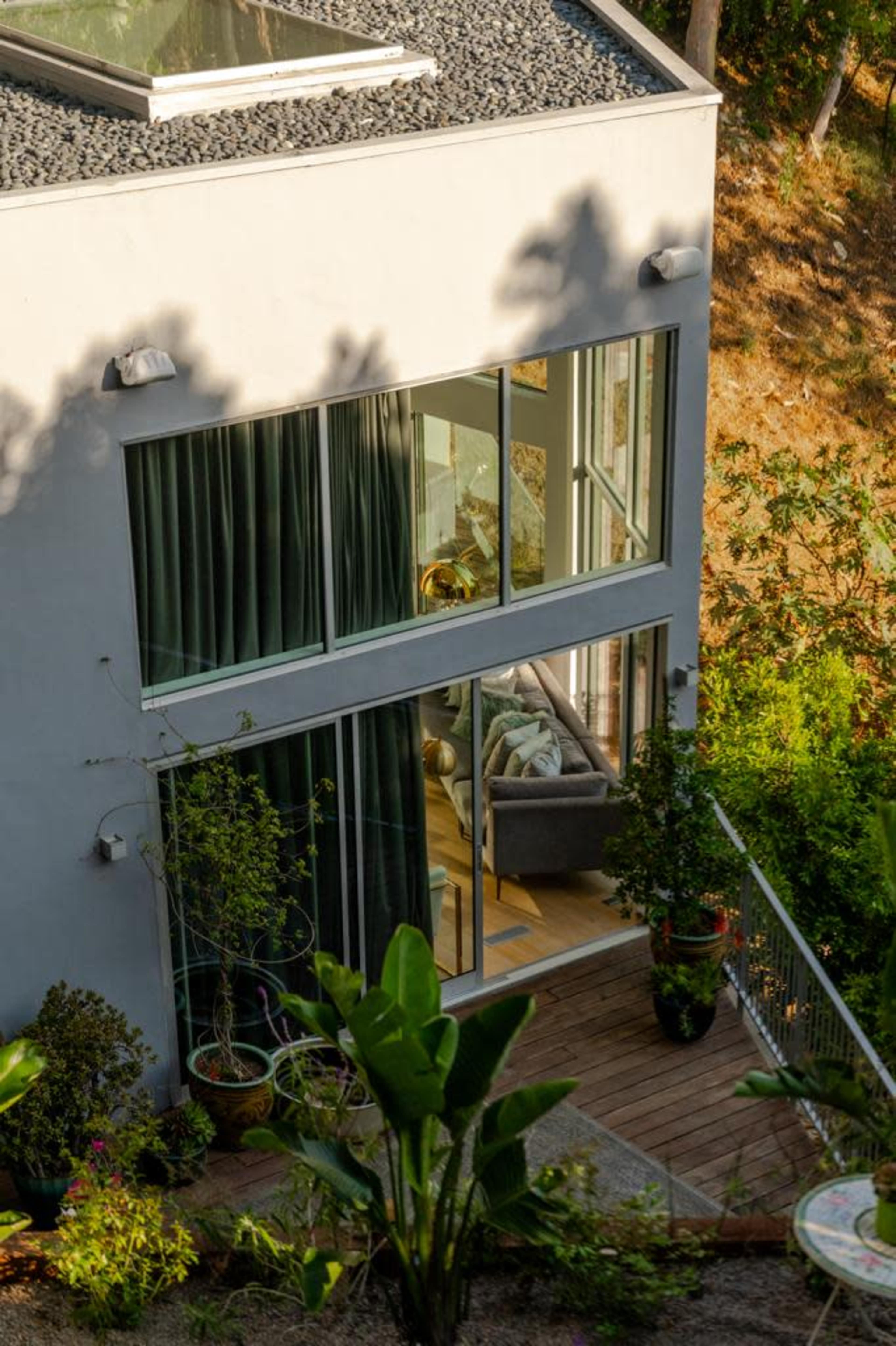 A modern house with large windows and a terrace surrounded by greenery is viewed from an elevated angle.