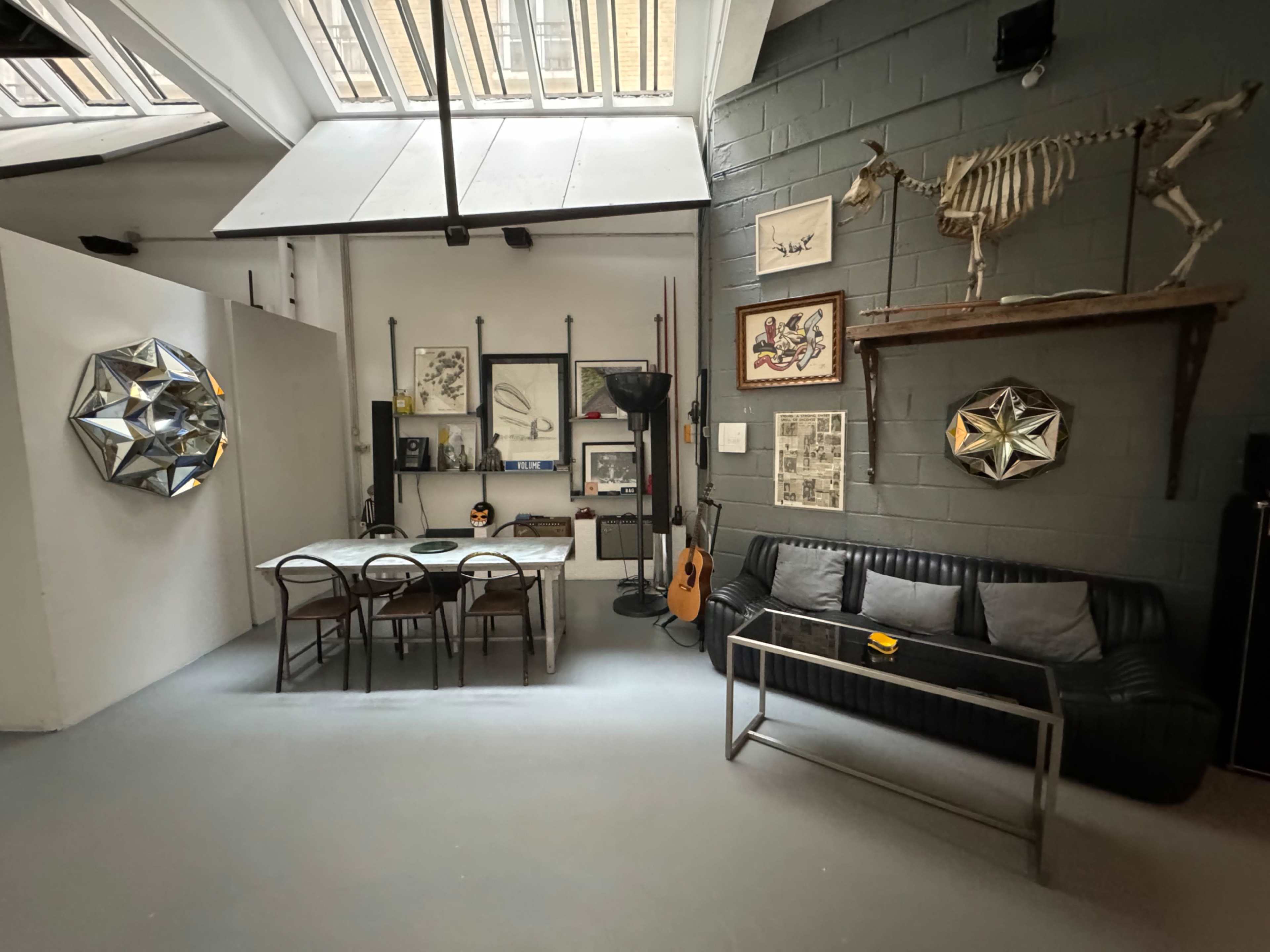 The image shows a modern interior space featuring a dining area with a table and chairs, a black sofa, and various artworks and decorations on the walls, including a sculpture and a guitar.
