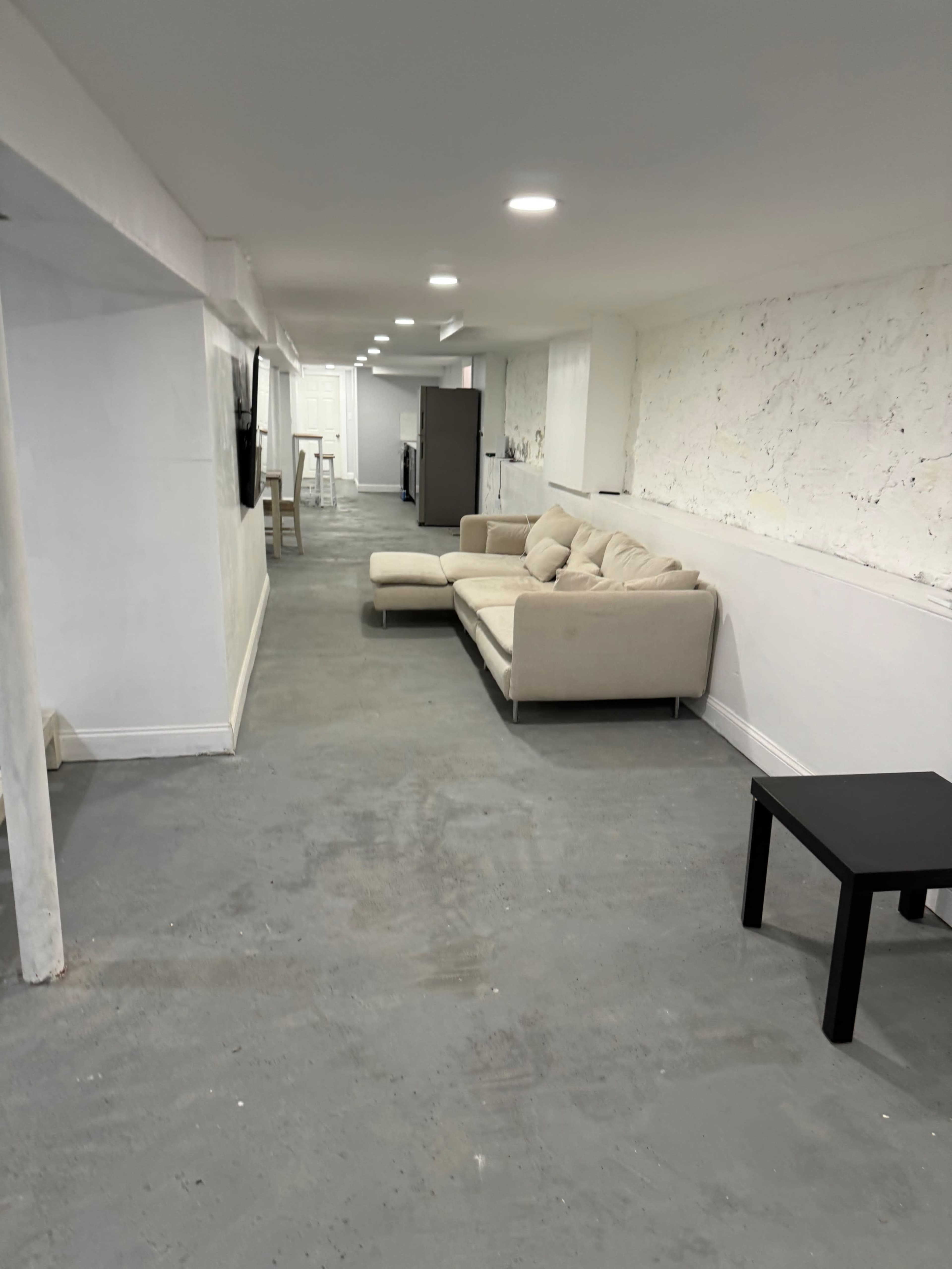 A long, narrow basement space featuring a light-colored sofa, a black table, and a refrigerator in the background.