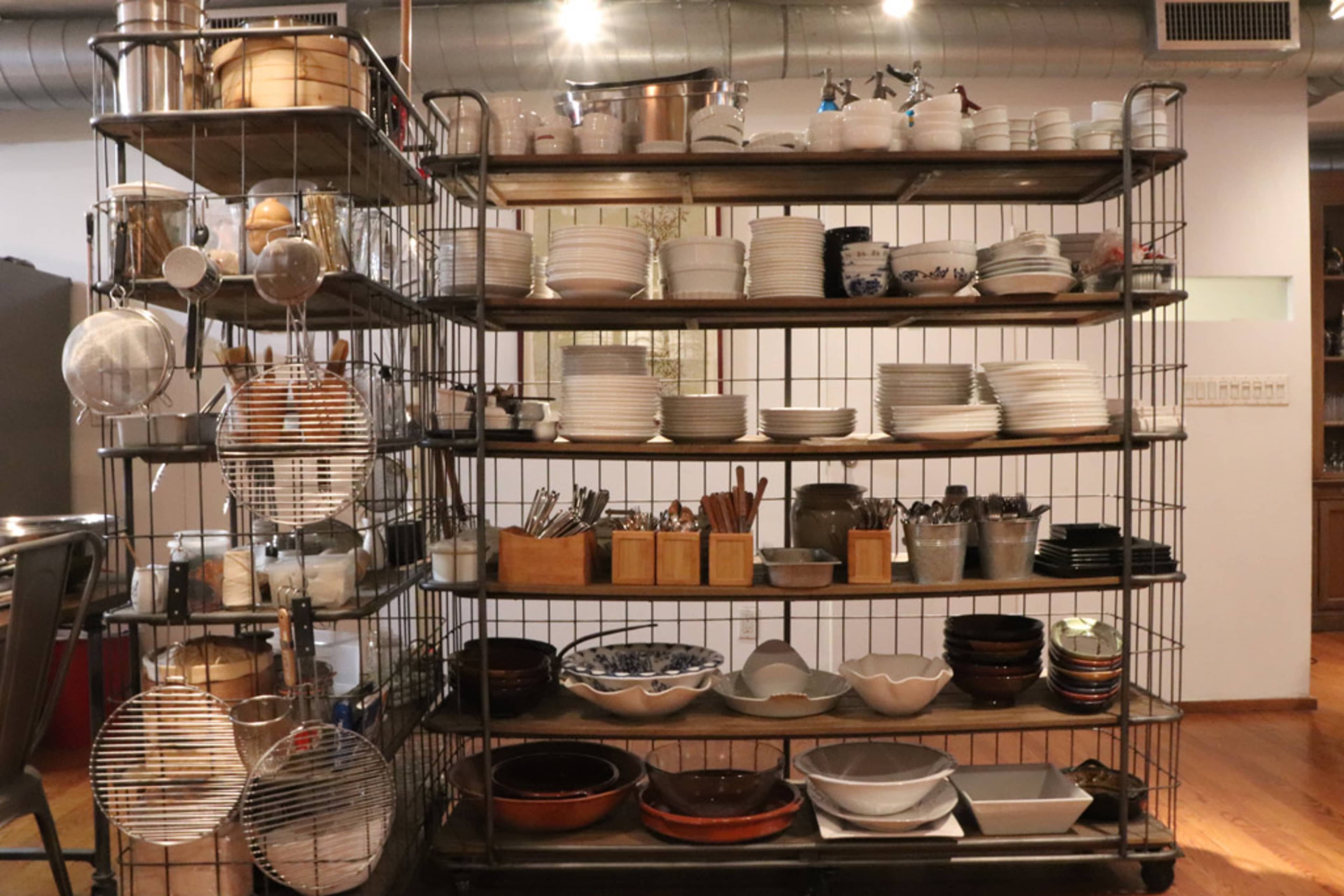 A metal shelving unit displays various kitchenware, including bowls, plates, utensils, and containers, organized across multiple shelves.