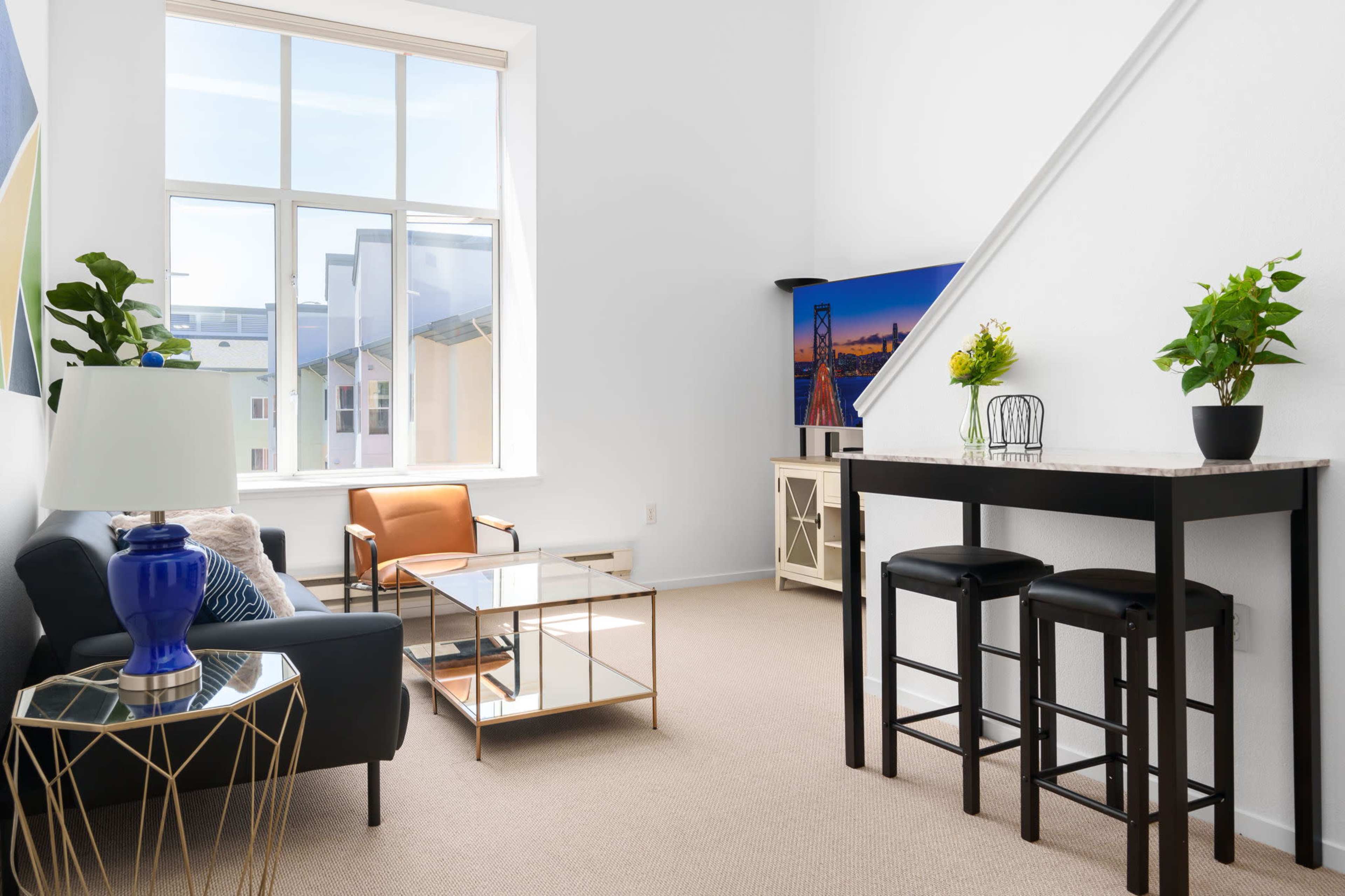 A bright, modern living space with a sofa, a coffee table, a bar table with stools, and a decorative plant near a large window.