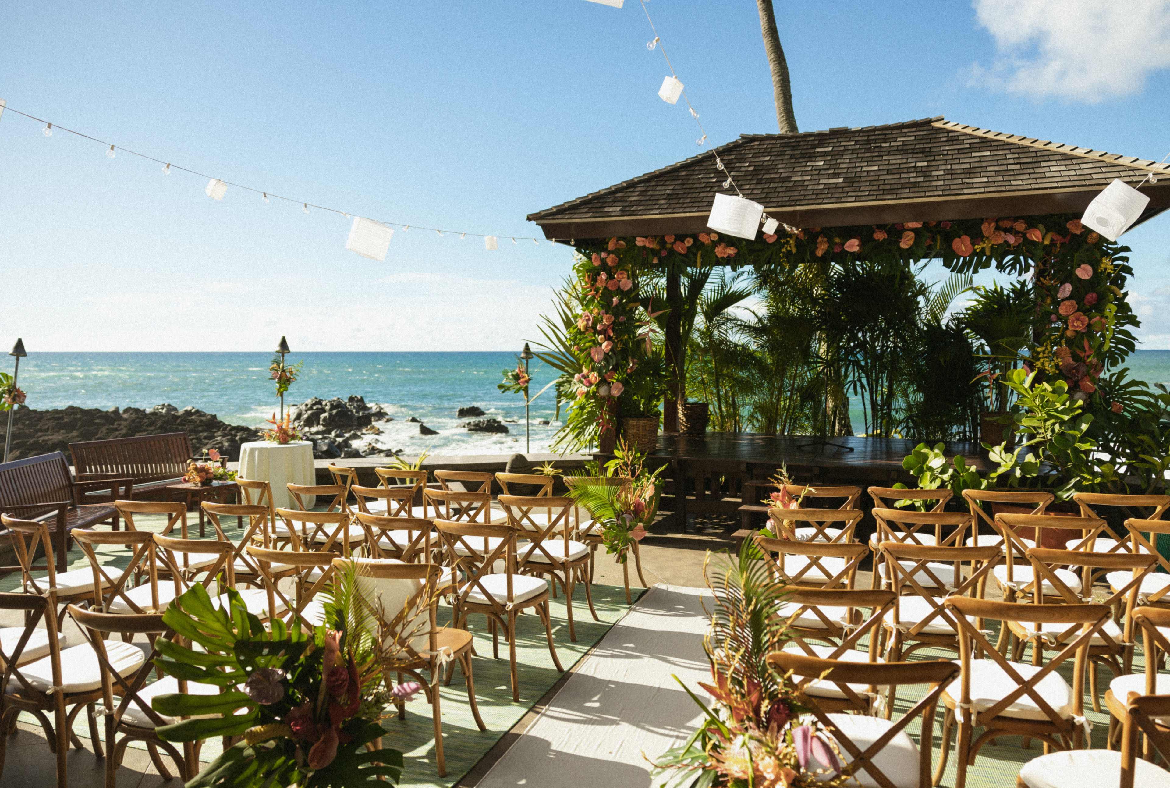 An outdoor wedding venue features rows of wooden chairs facing a decorated pavilion with a view of the ocean.