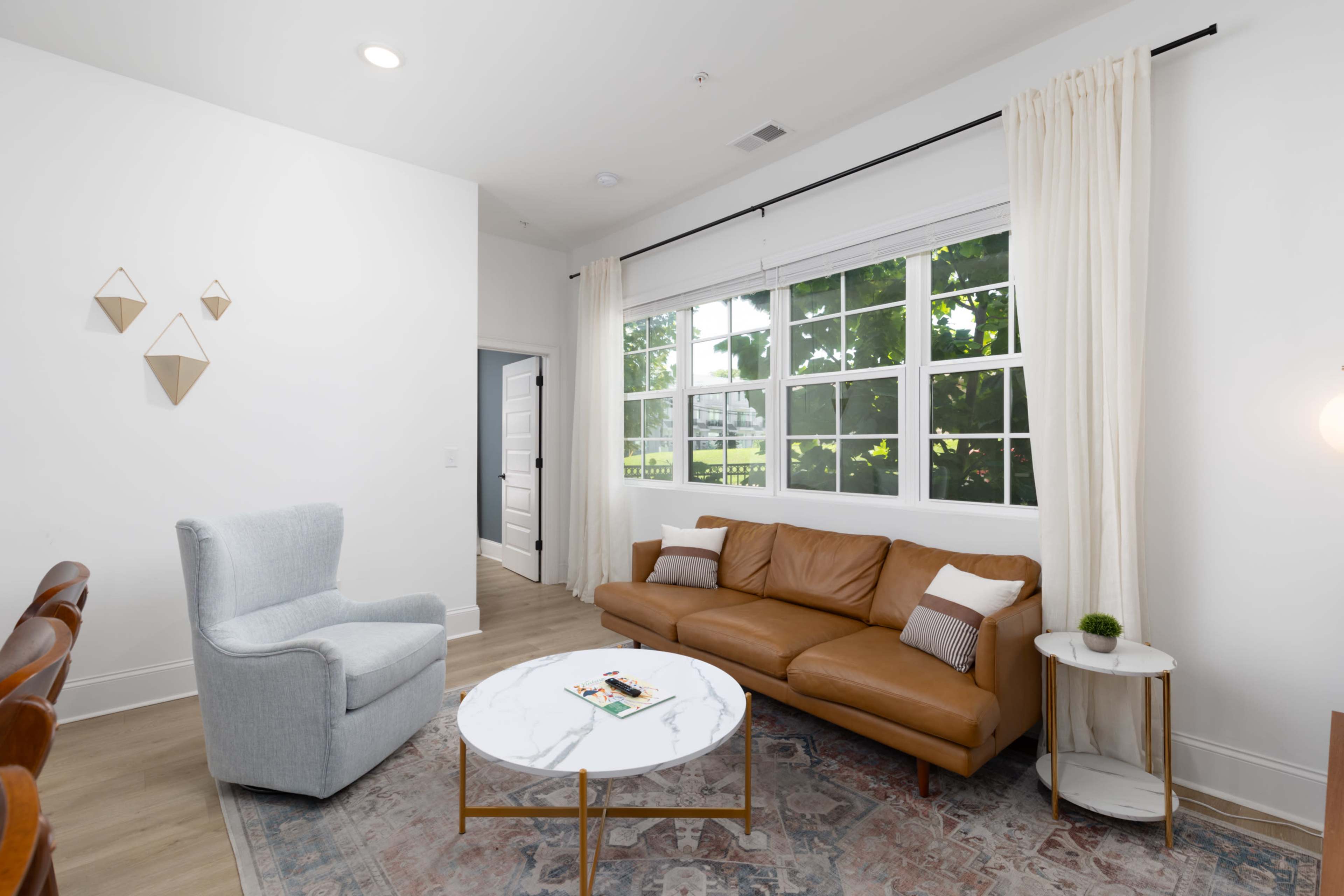 The image shows a living room with a brown leather sofa, a round marble coffee table, and large windows that let in natural light.