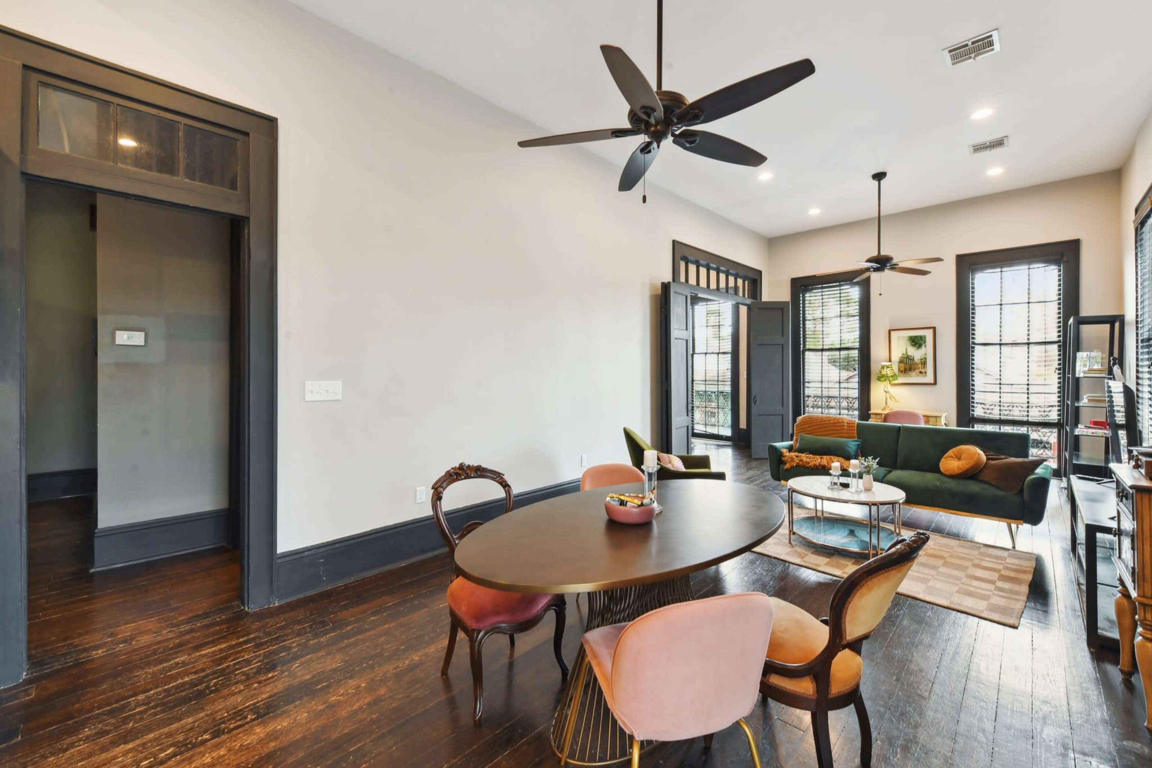 The scene shows a modern living space featuring a round dining table with colorful chairs, a green sofa, and large windows with blinds.