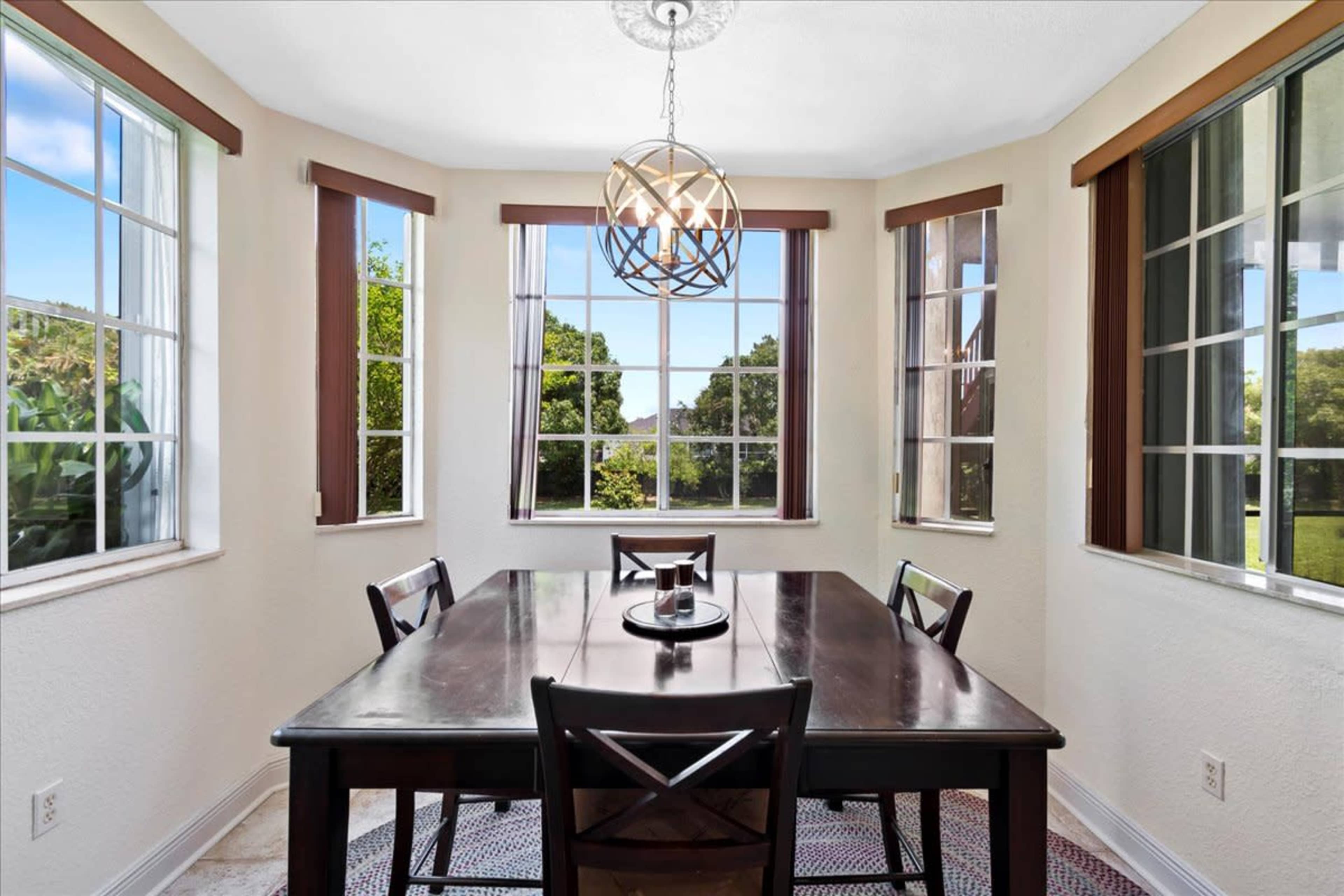 A dining area features a dark wooden table surrounded by black chairs, illuminated by a geometric light fixture, with windows showing green foliage outside.