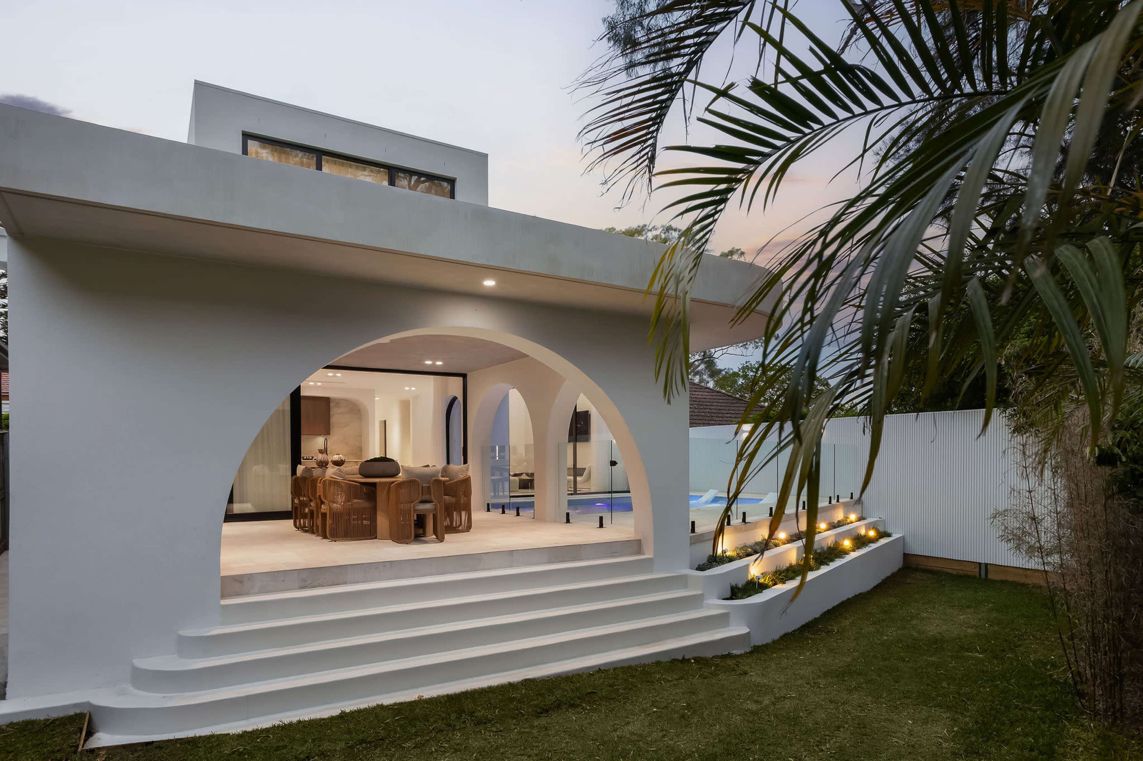 The image shows a modern white house with arched features, a patio area with outdoor dining furniture, and a swimming pool visible in the background.