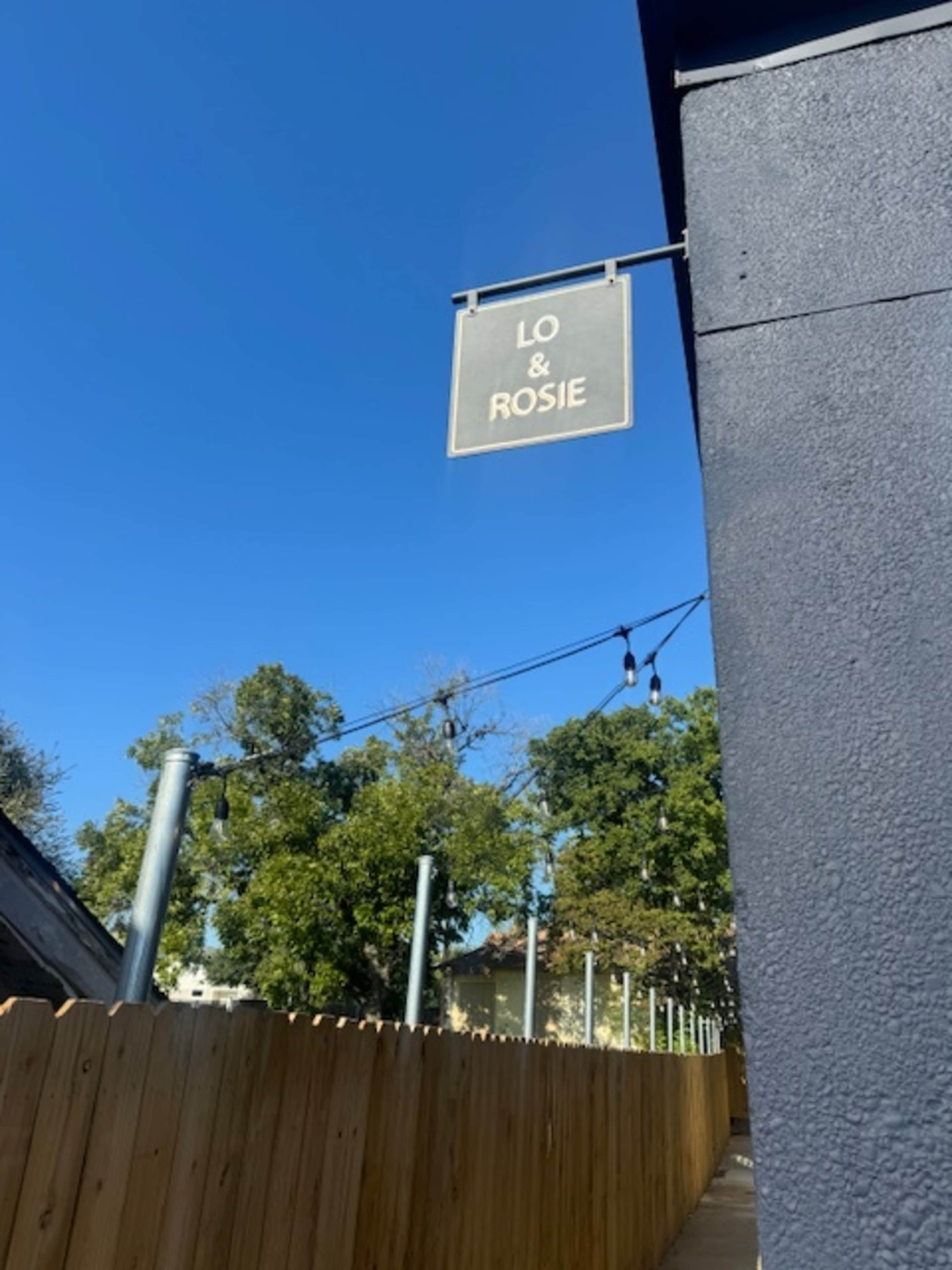 A sign reading "LO & ROSIE" hangs from a building near a wooden fence, with clear blue skies and trees in the background.