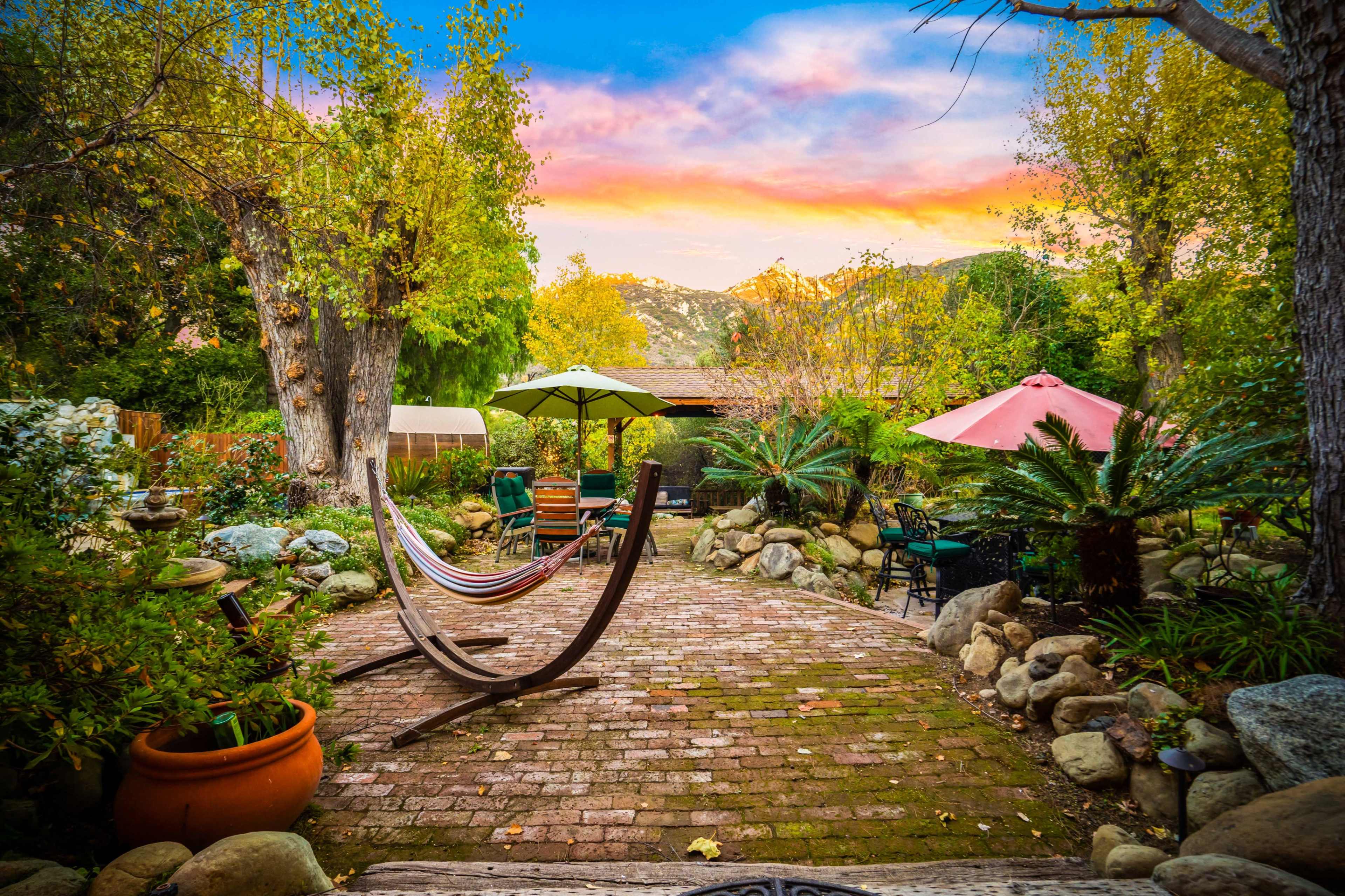 A brick pathway leads through a landscaped garden featuring a hammock, patio furniture, and varied greenery under a colorful sky.